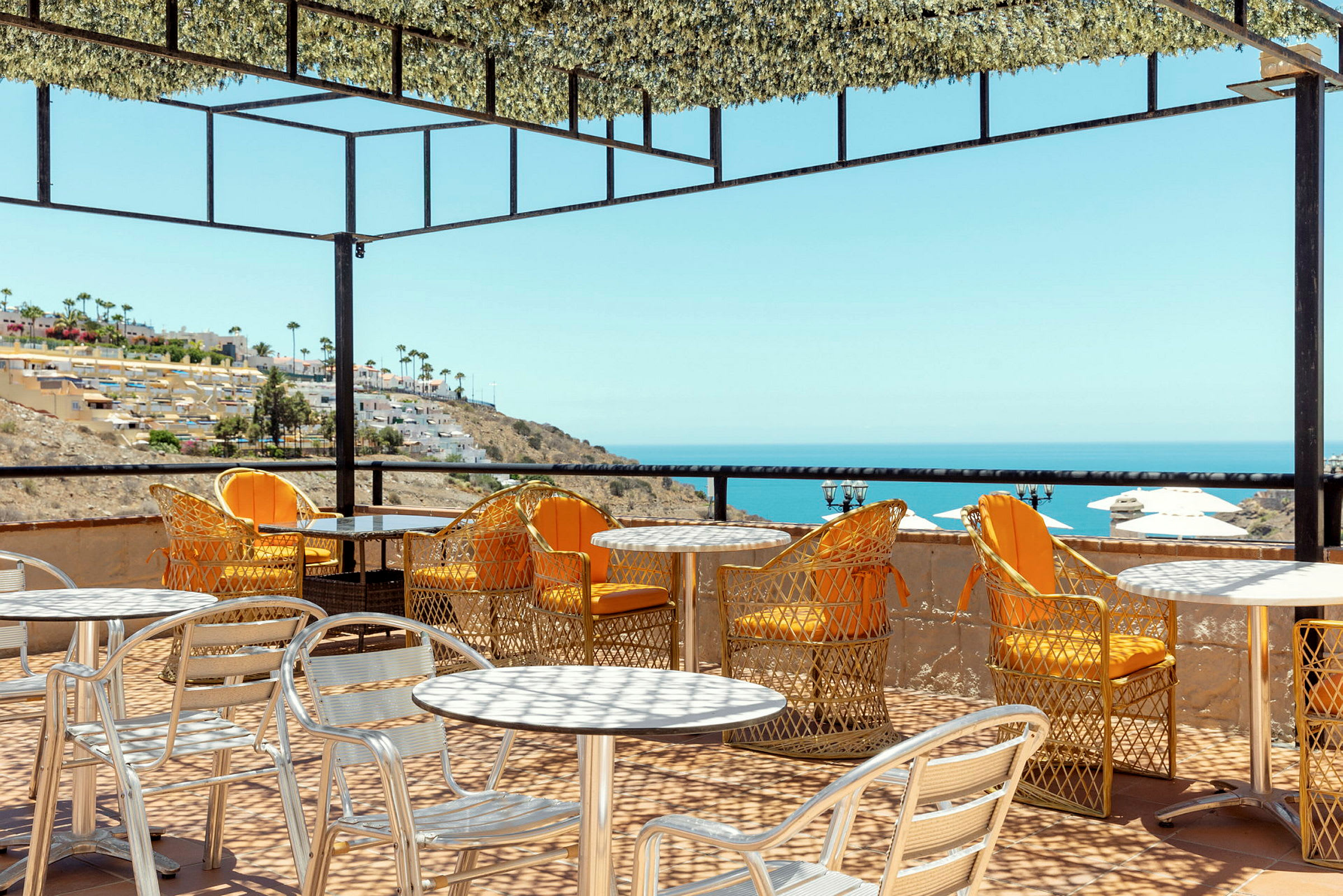 Udendørs servering med borde og stole på en terrasse med udsigt over havet og en skråning med bygninger. (AI-genereret tekst)