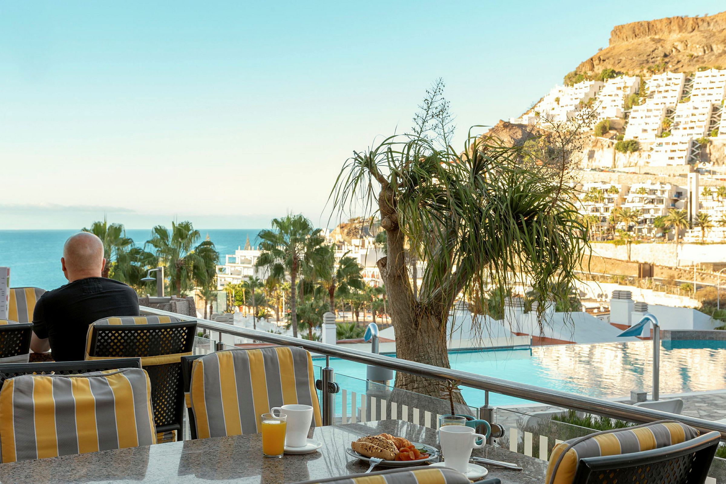 En person sidder på en terrasse med udsigt over havet, træer og bygninger, med en kop kaffe og morgenmad foran sig. (AI-genereret tekst)