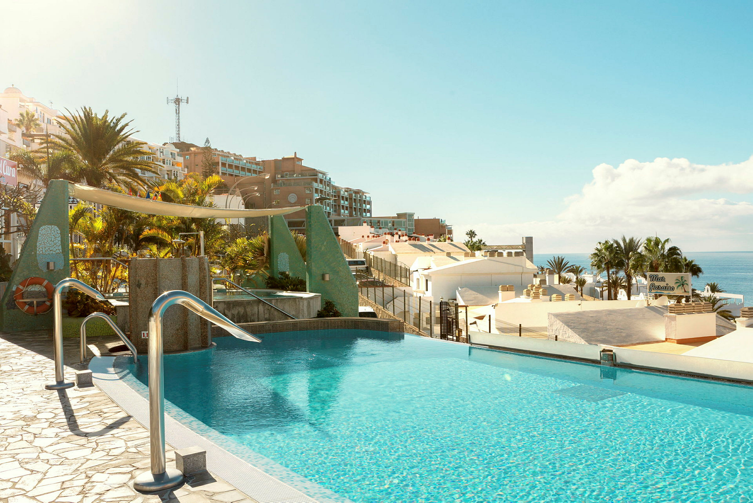En infinity-pool med udsigt over hustage, palmer og havet i et solrigt landskab. (AI-genereret tekst)