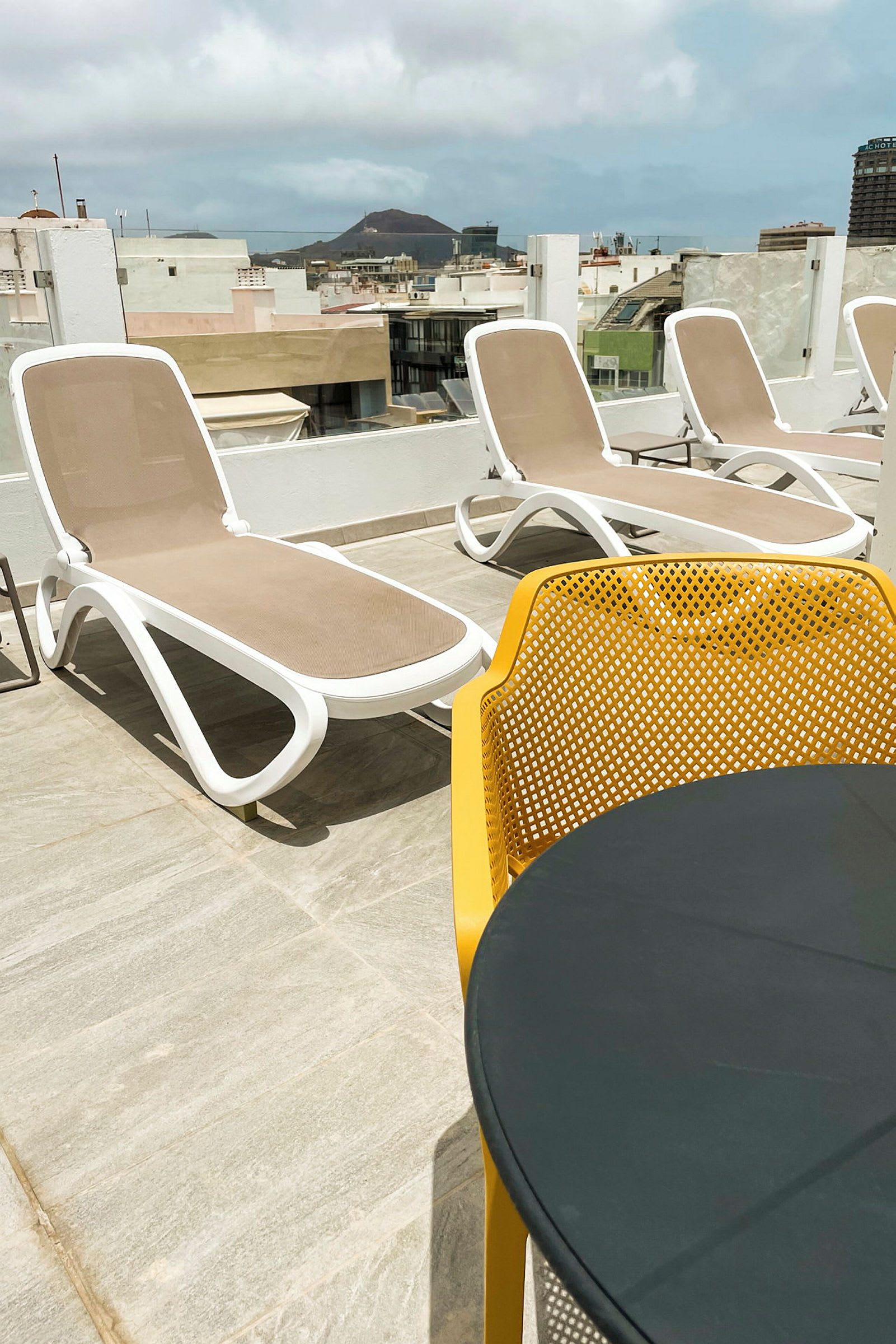 En solterrasse med liggestole og et bord med gule stole i forgrunden, byudsigt og et bjerg i baggrunden. (AI-genereret tekst)
