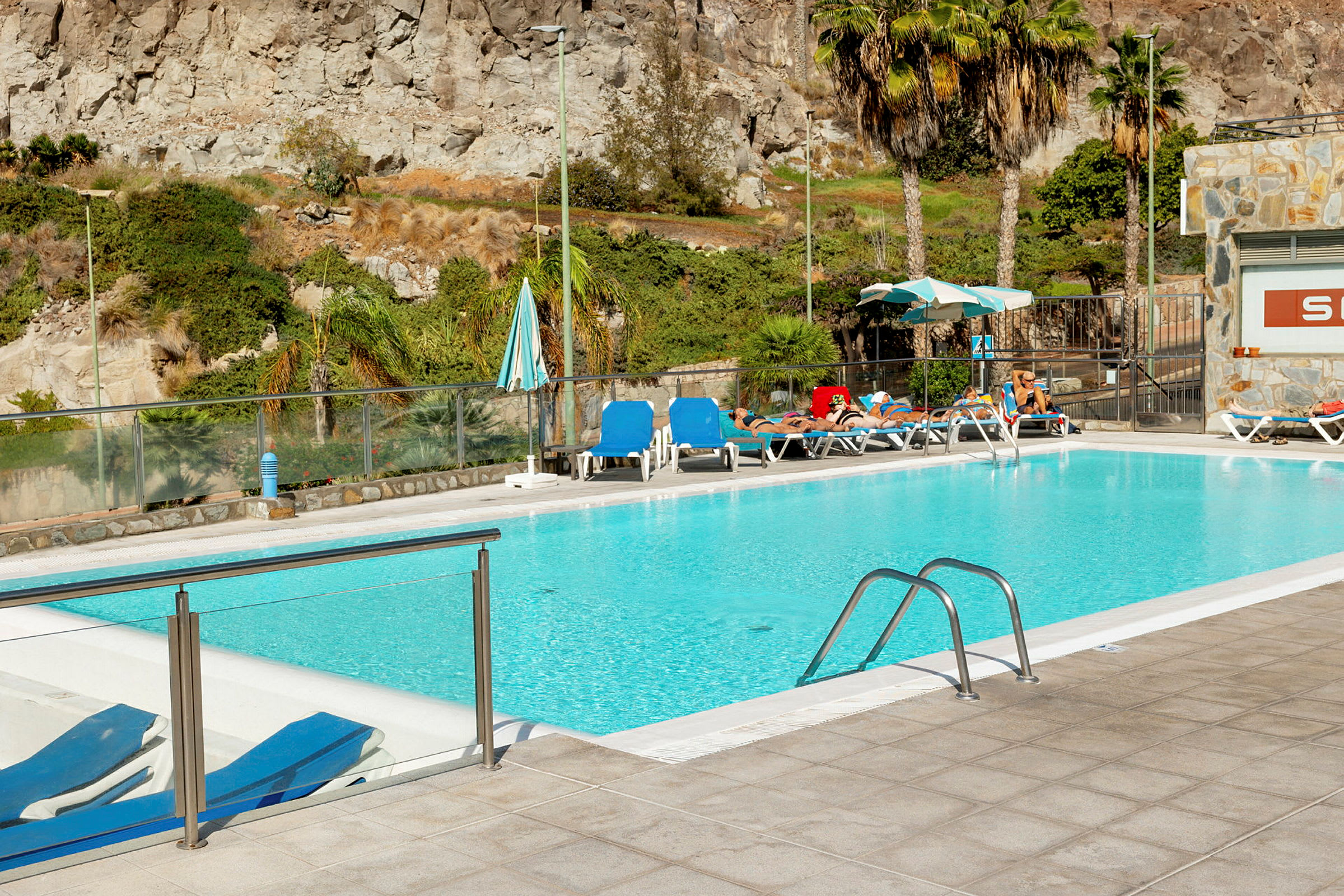 En swimmingpool omgivet af liggestole og parasoller, med klipper og vegetation i baggrunden. (AI-genereret tekst)