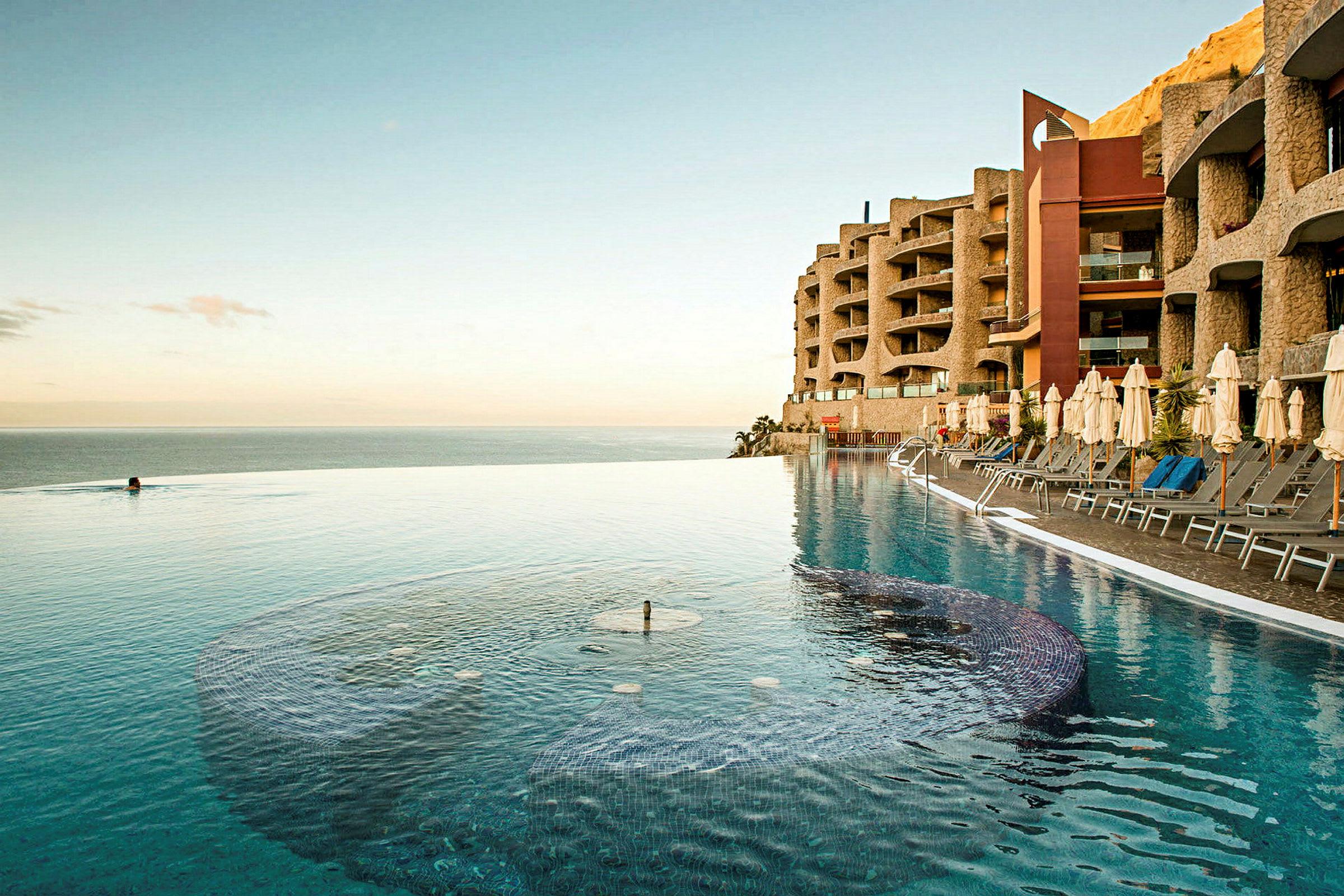 En infinitypool ved havet med liggestole og parasoller på siden og et hotel med moderne arkitektur i baggrunden. (AI-genereret tekst)