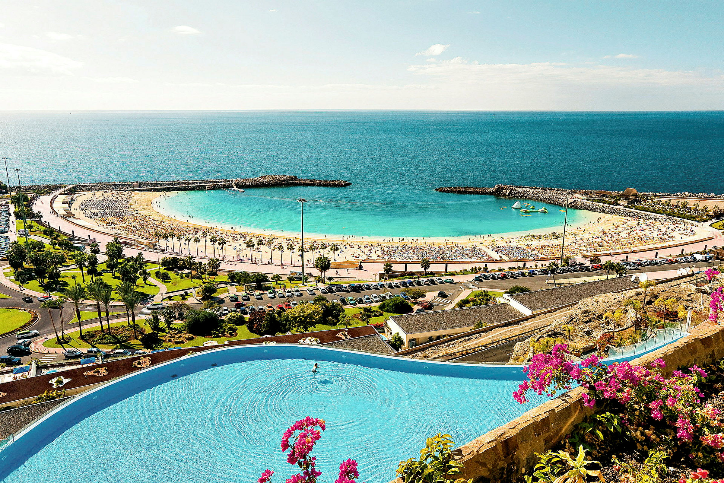 En strand med gul sand og klart blåt hav, omgivet af en beskyttende mole og grønne områder. En pool med blomster i forgrunden. (AI-genereret tekst)