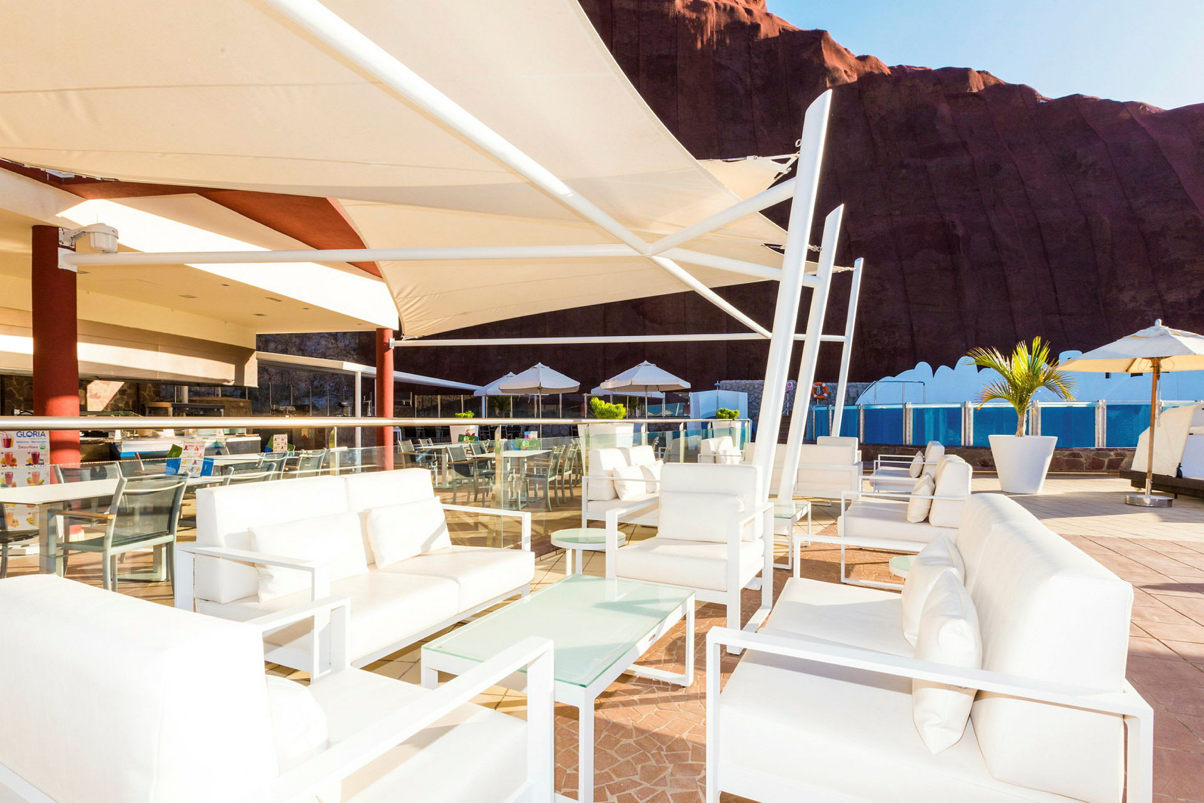 En udendørs terrasse med hvide møbler, solskærme og udsigt til en pool. (AI-genereret tekst)