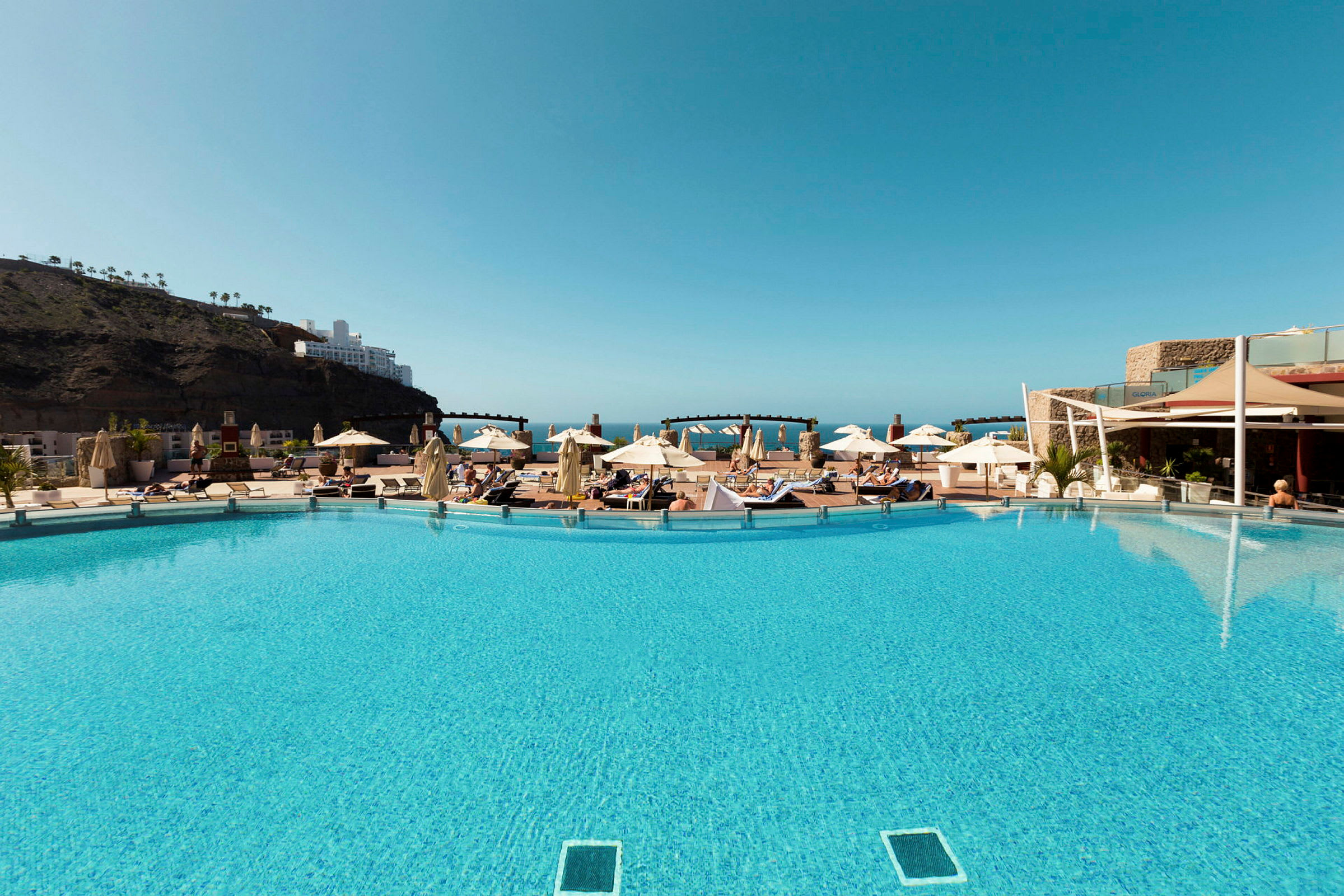 En swimmingpool med liggestole og parasoller, omgivet af bygninger og bjerge under en klar blå himmel. (AI-genereret tekst)