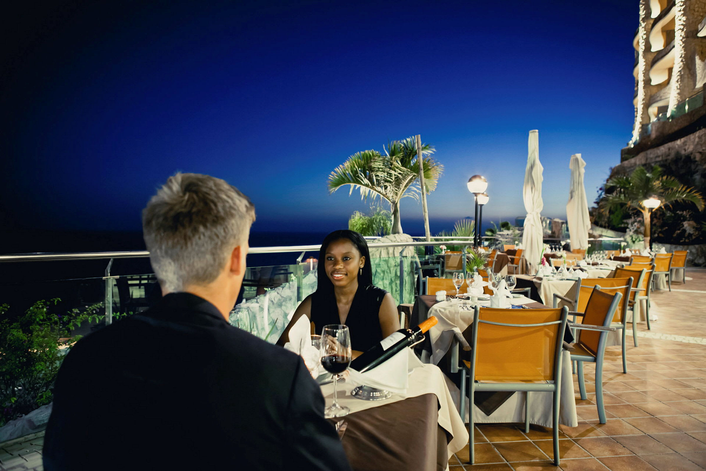 To personer ved et bord på en restaurant med udsigt over havet og tropiske planter i aftentimen. (AI-genereret tekst)