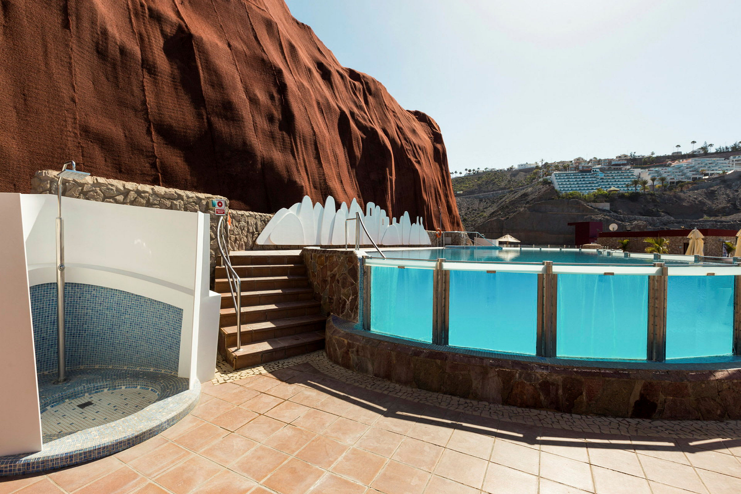 En pool med klart blåt vand omgivet af en stenmur og terrasse. Bag poolen ses en stor rød klippe og hvide bygninger. (AI-genereret tekst)