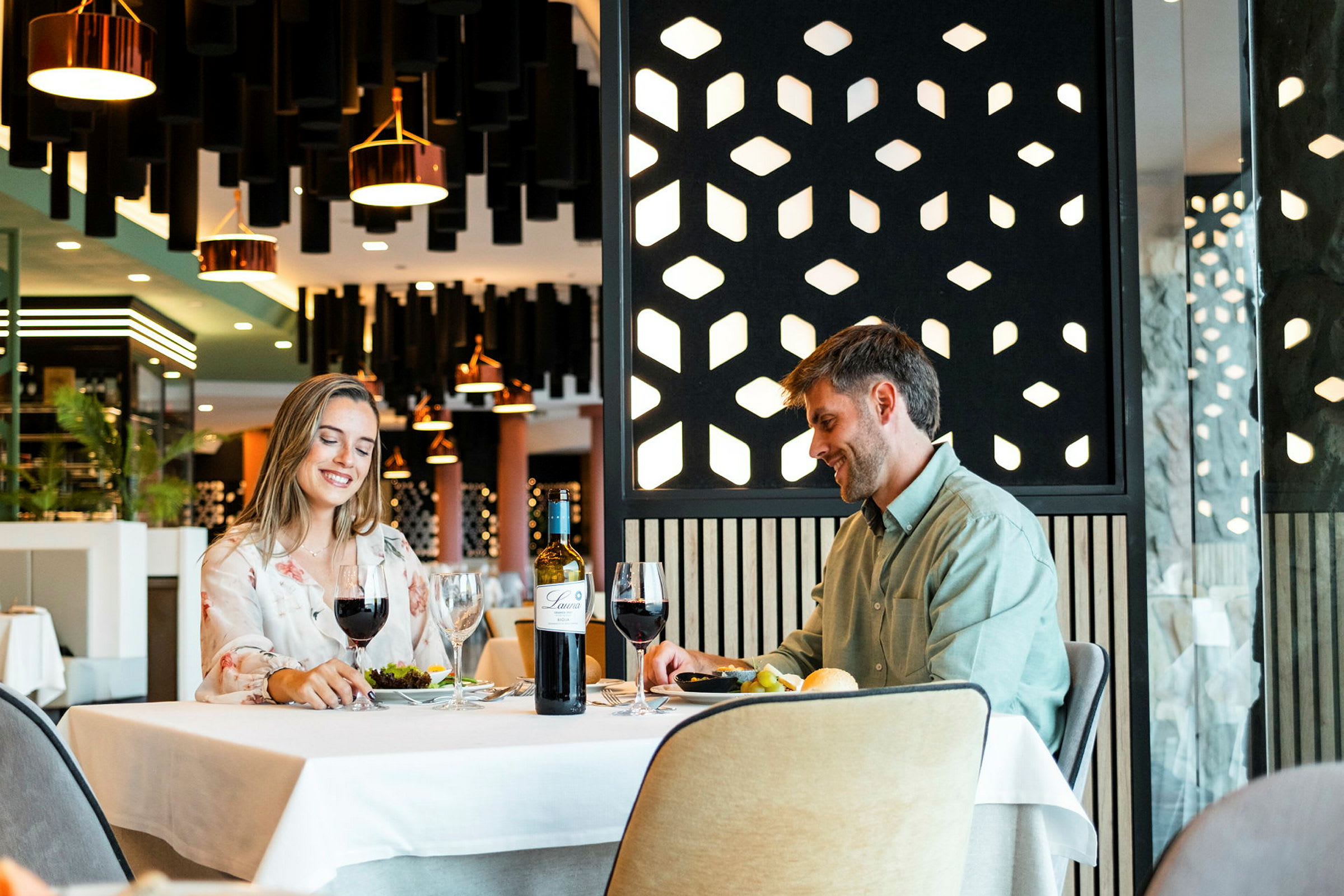 To personer sidder ved et bord på en restaurant med moderne indretning. På bordet er der glas, en vinflaske og tallerkener med mad. (AI-genereret tekst)