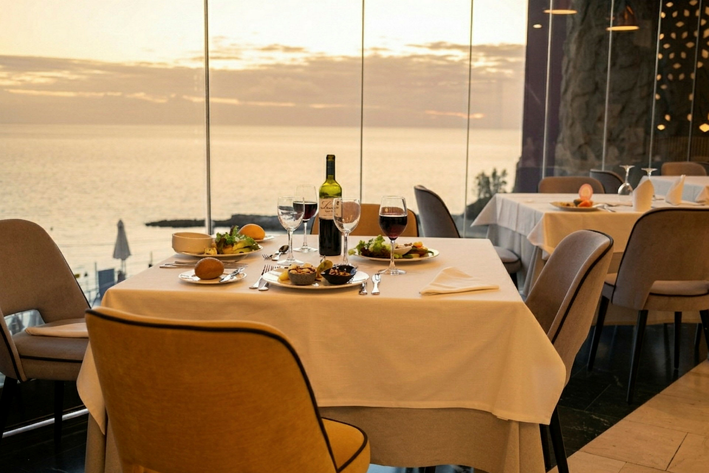 Et bord på en restaurant med hvide duge, mad og vin placeret på bordet, med havudsigt gennem store vinduer. (AI-genereret tekst)