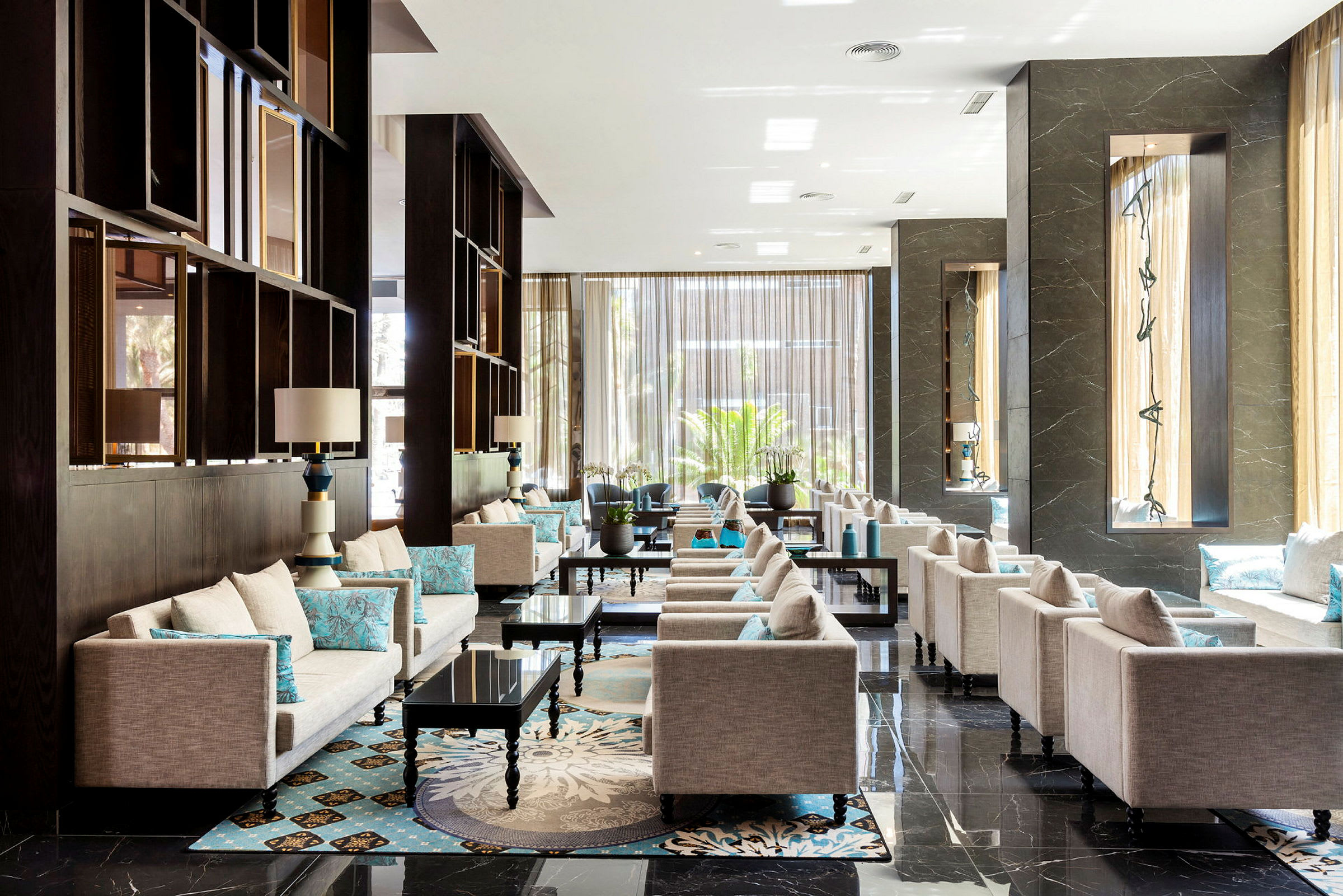 En elegant hotellobby med beige sofaer og turkise puder, mørke borde og store vinduer, der lukker naturligt lys ind. (AI-genereret tekst)