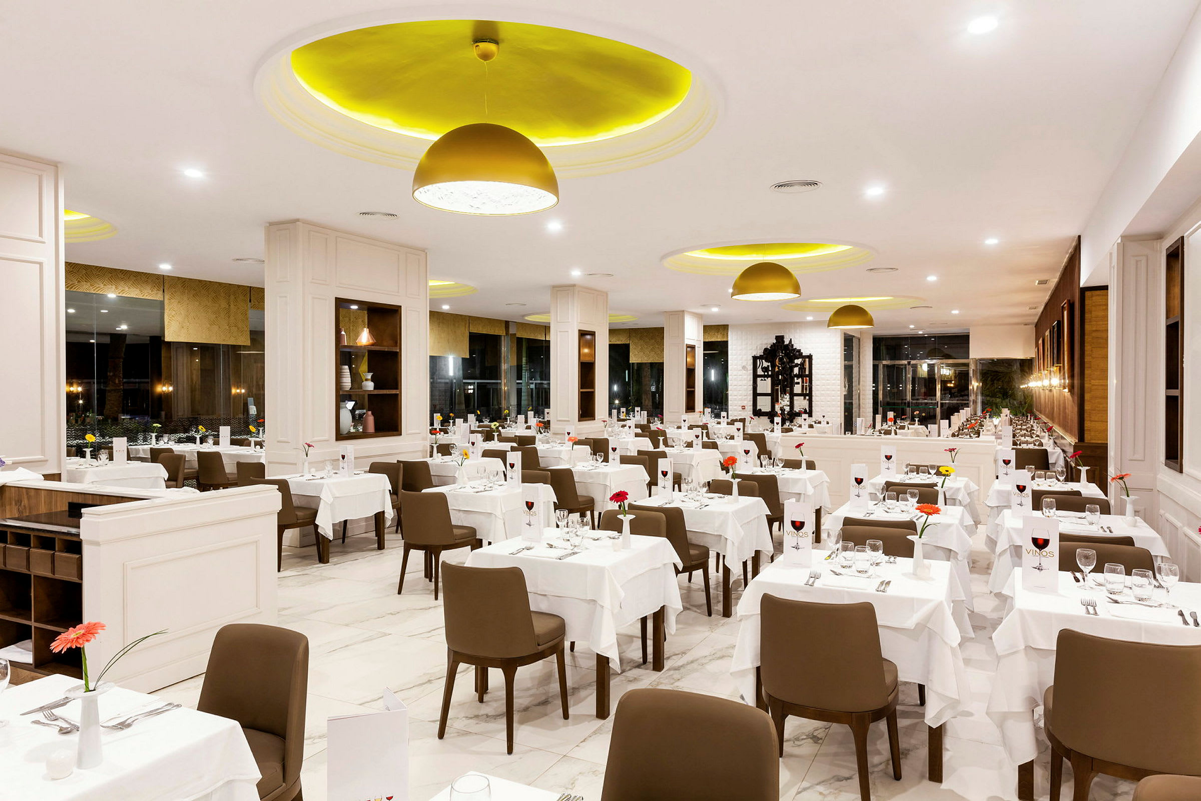 Et interiørbillede af en lys og elegant restaurant med mange borde dækket med hvide duge, brune stole og dekorationer. (AI-genereret tekst)