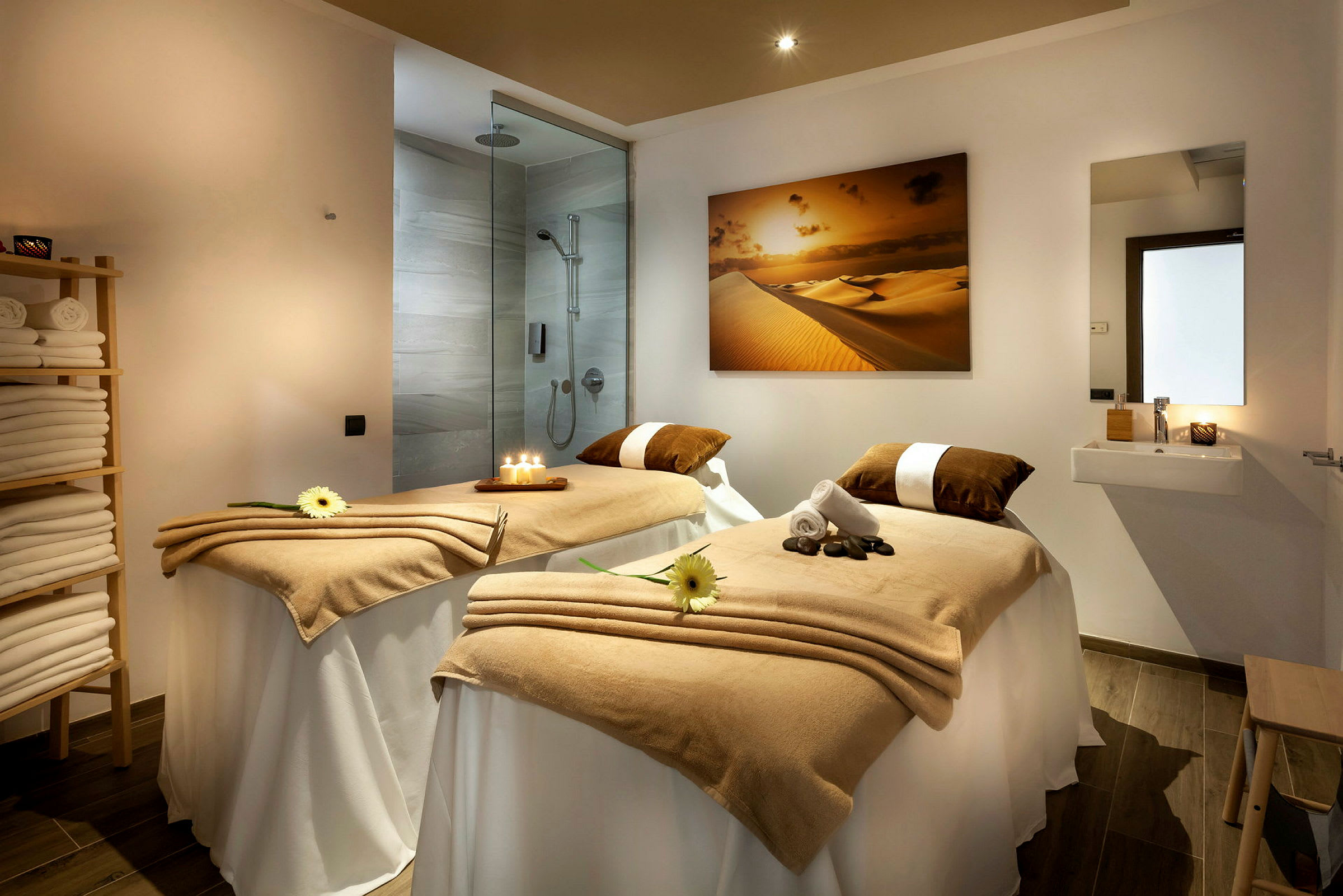 Et behandlingsrum i et spa med to massageborde dækket med beige tæpper, stearinlys og dekorative blomster. En bruser og håndvask ses i baggrunden. (AI-genereret tekst)