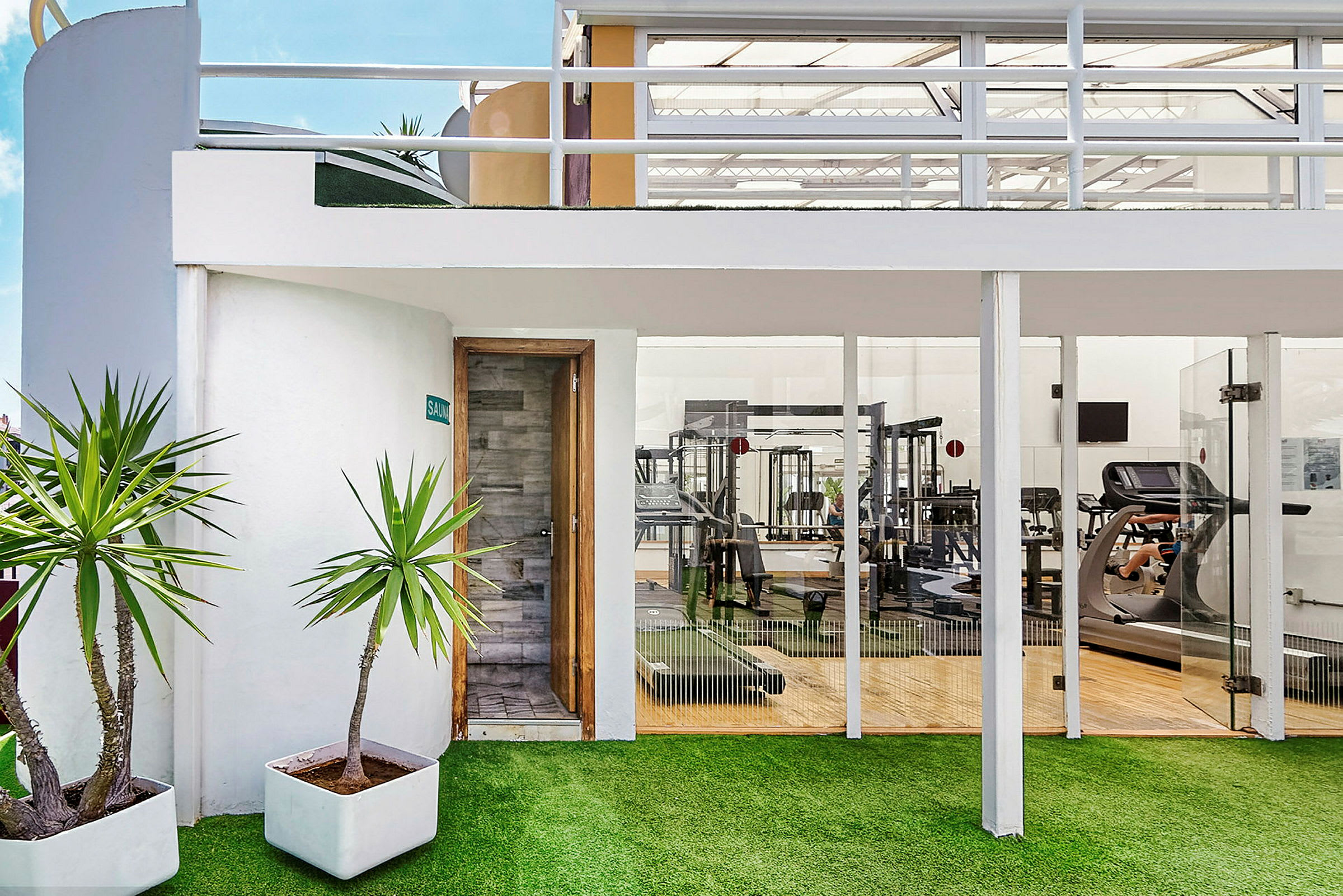 Udvendigt af et fitnesscenter med glasvægge og en saunadør, samt grønne planter i potter foran. (AI-genereret tekst)