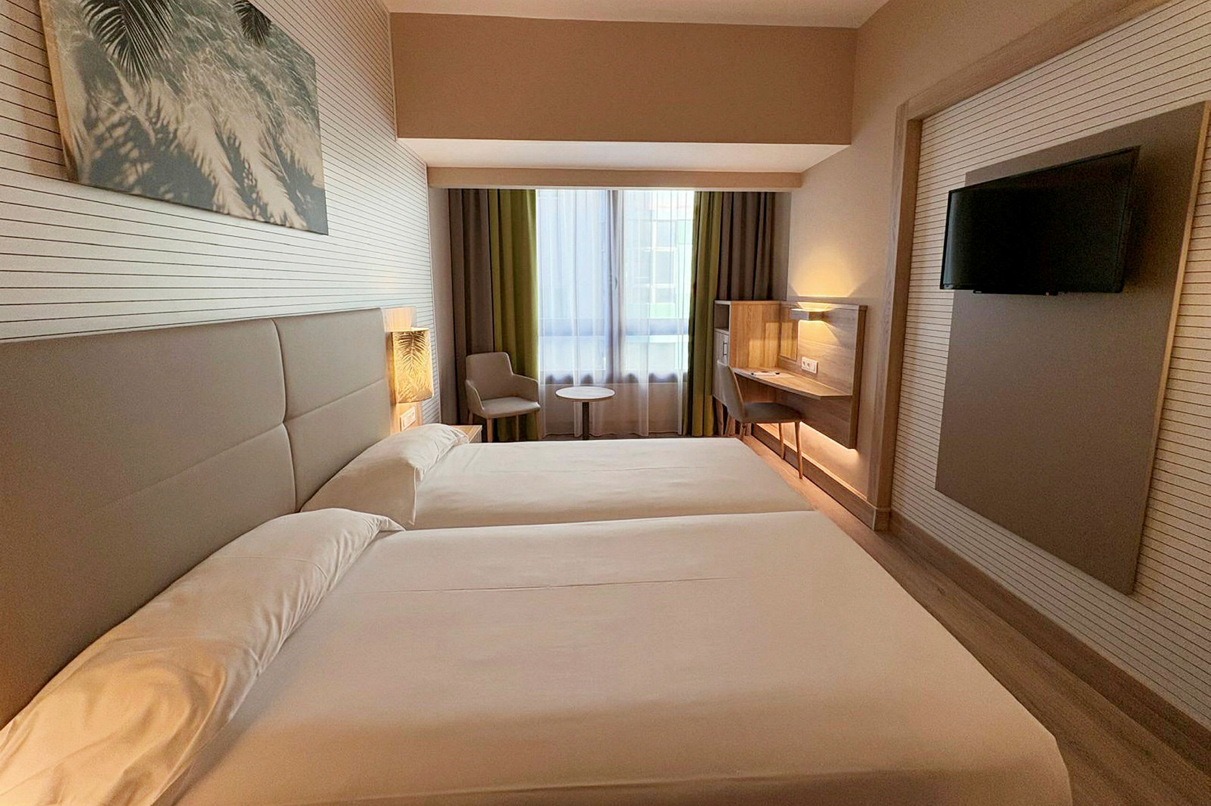 Hotelværelse med to enkeltsenge, et vindue med gardiner, siddeområde med bord og et vægmonteret fladskærms-TV. (AI-genereret tekst)