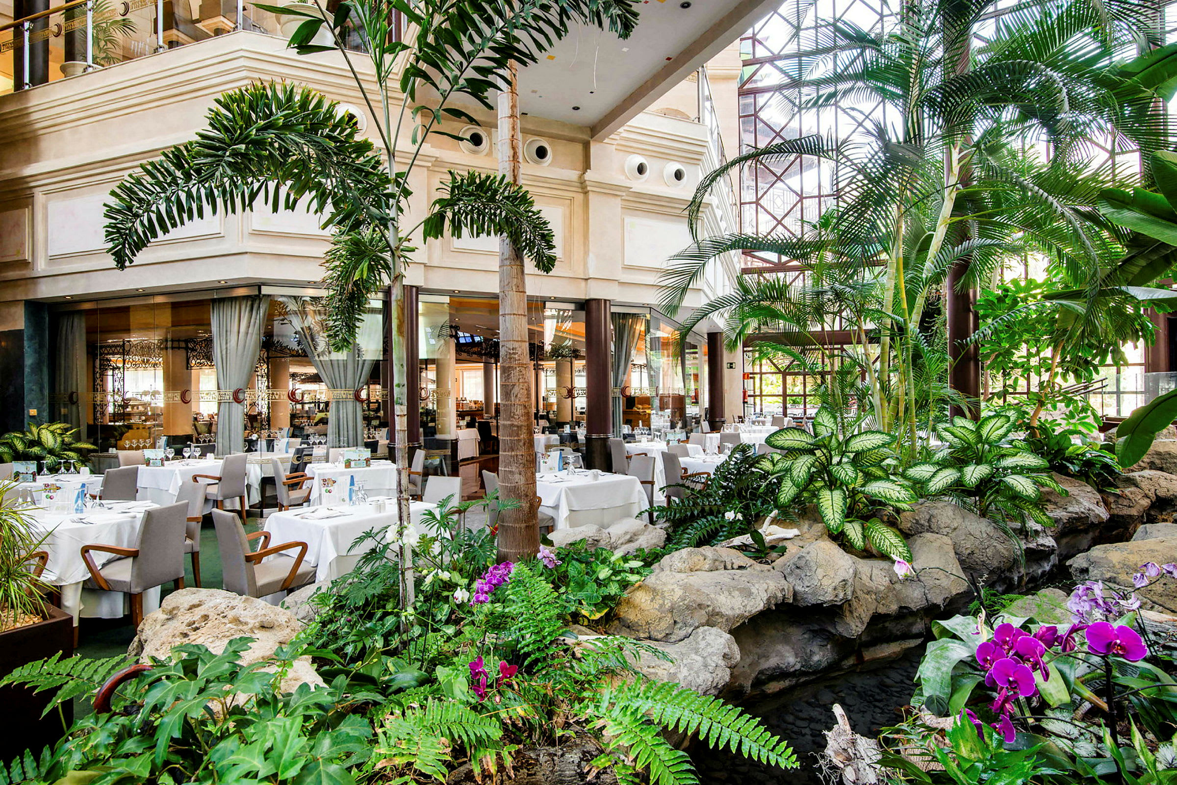 En restaurant med hvide duge og siddepladser er omgivet af frodig vegetation og sten. (AI-genereret tekst)