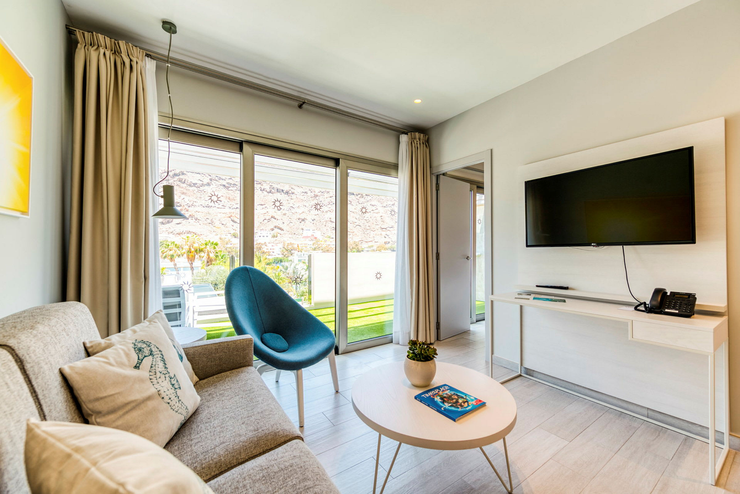 Et moderne hotelværelse med sofa, lænestol og vægmonteret TV samt glasdøre, der fører ud til en terrasse. (AI-genereret tekst)
