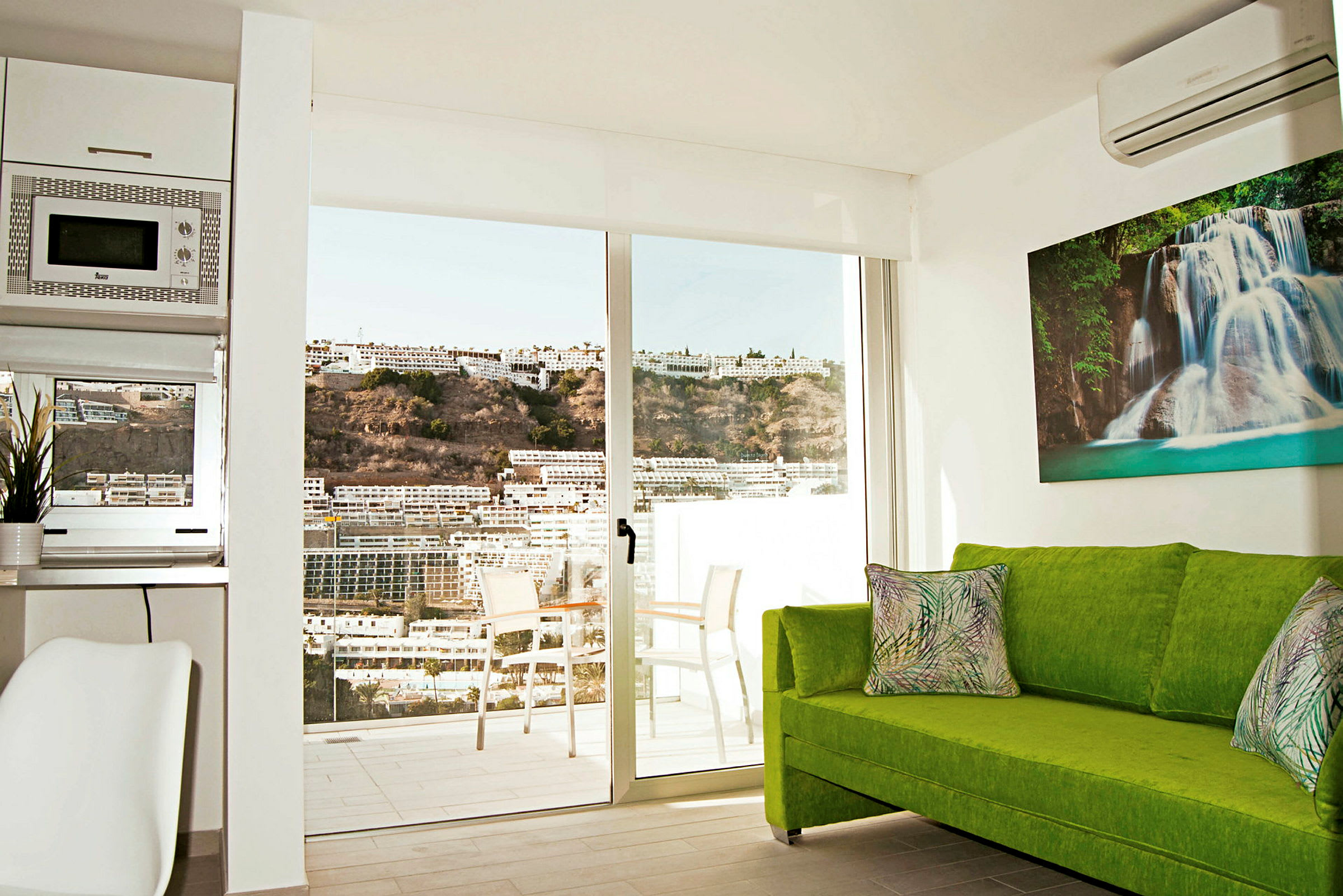 Interiør af et værelse med en grøn sofa, en glasdør mod en balkon og udsigt over bygninger og en bakke. (AI-genereret tekst)