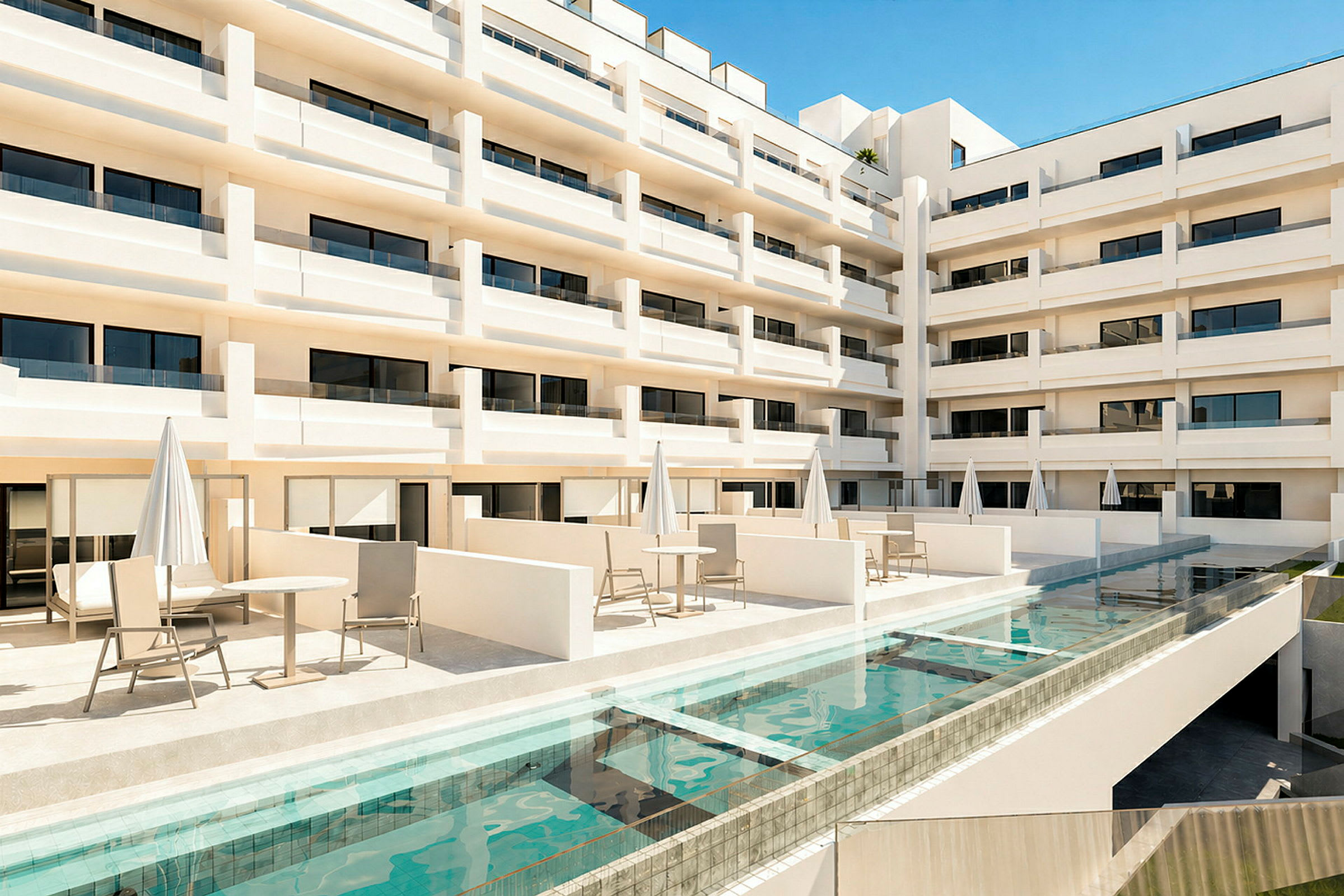 Udsigt over en moderne hotelbygning med flere etager og altaner. Foran bygningen findes en terrasse med borde, stole og parasoller samt en swimmingpool. (AI-genereret tekst)