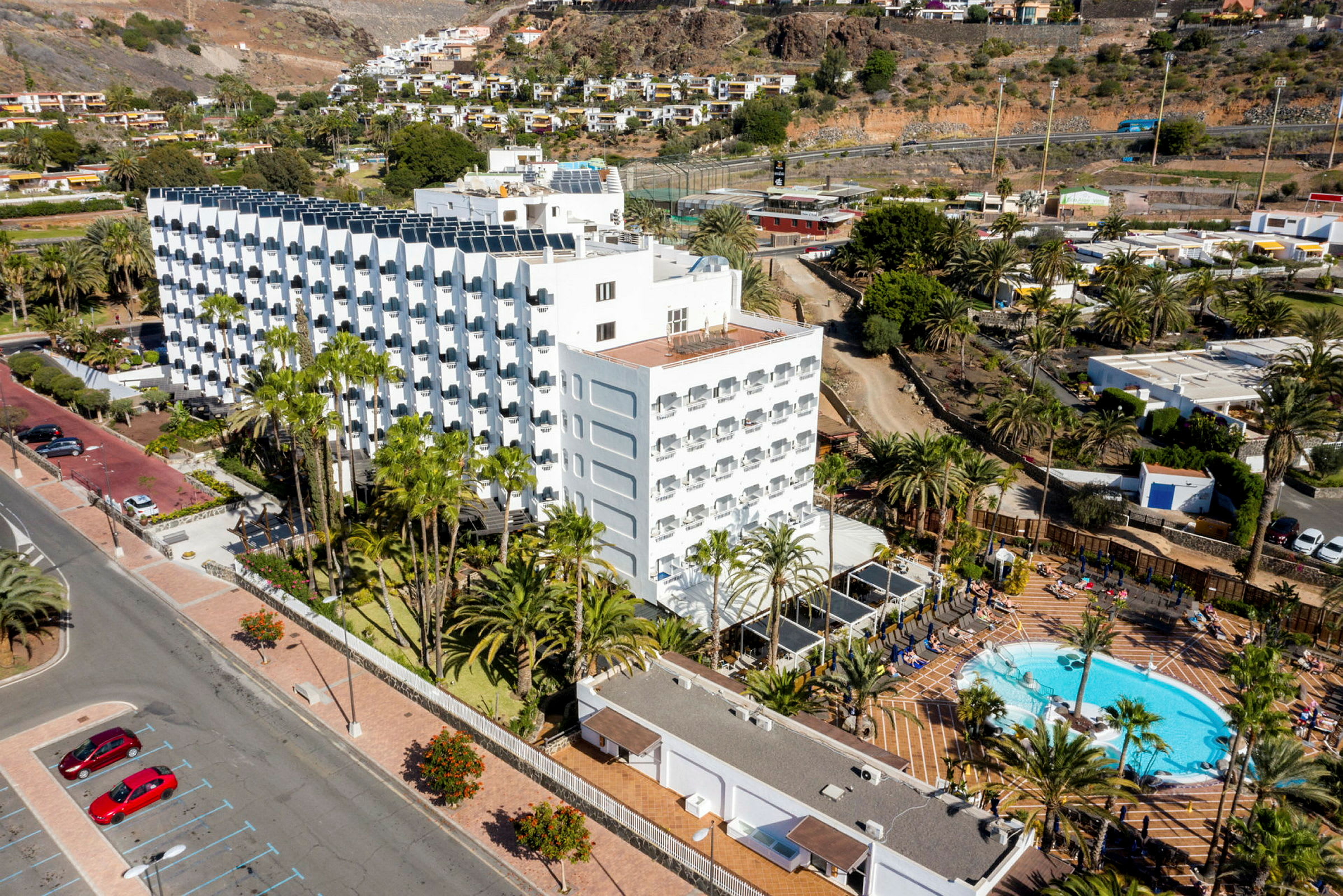 Et hotelkompleks med pool og palmetræer omgivet af bjerge og grønne områder. (AI-genereret tekst)