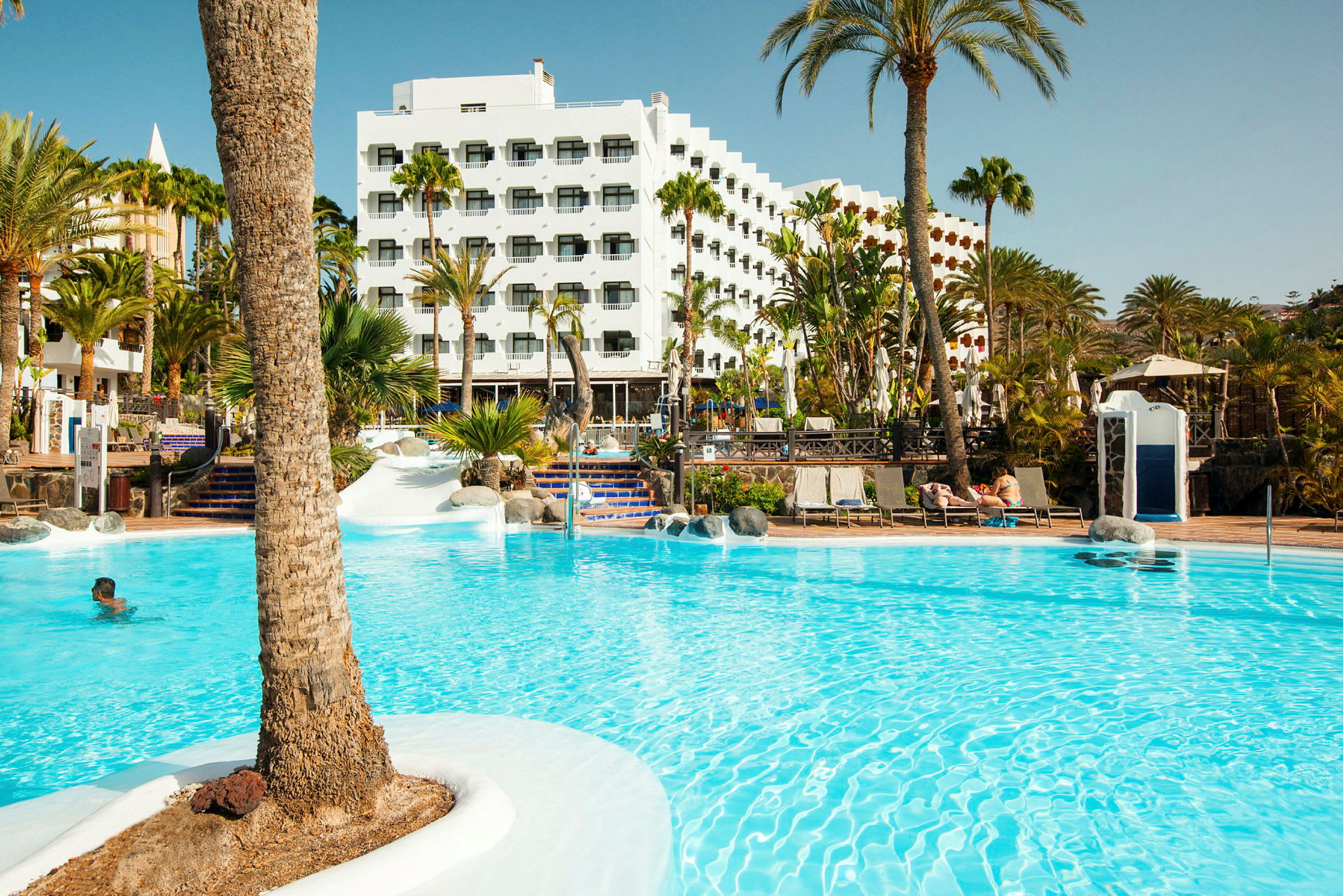 Billede af en udendørs pool med palmer og hotelbygninger i baggrunden. (AI-genereret tekst)