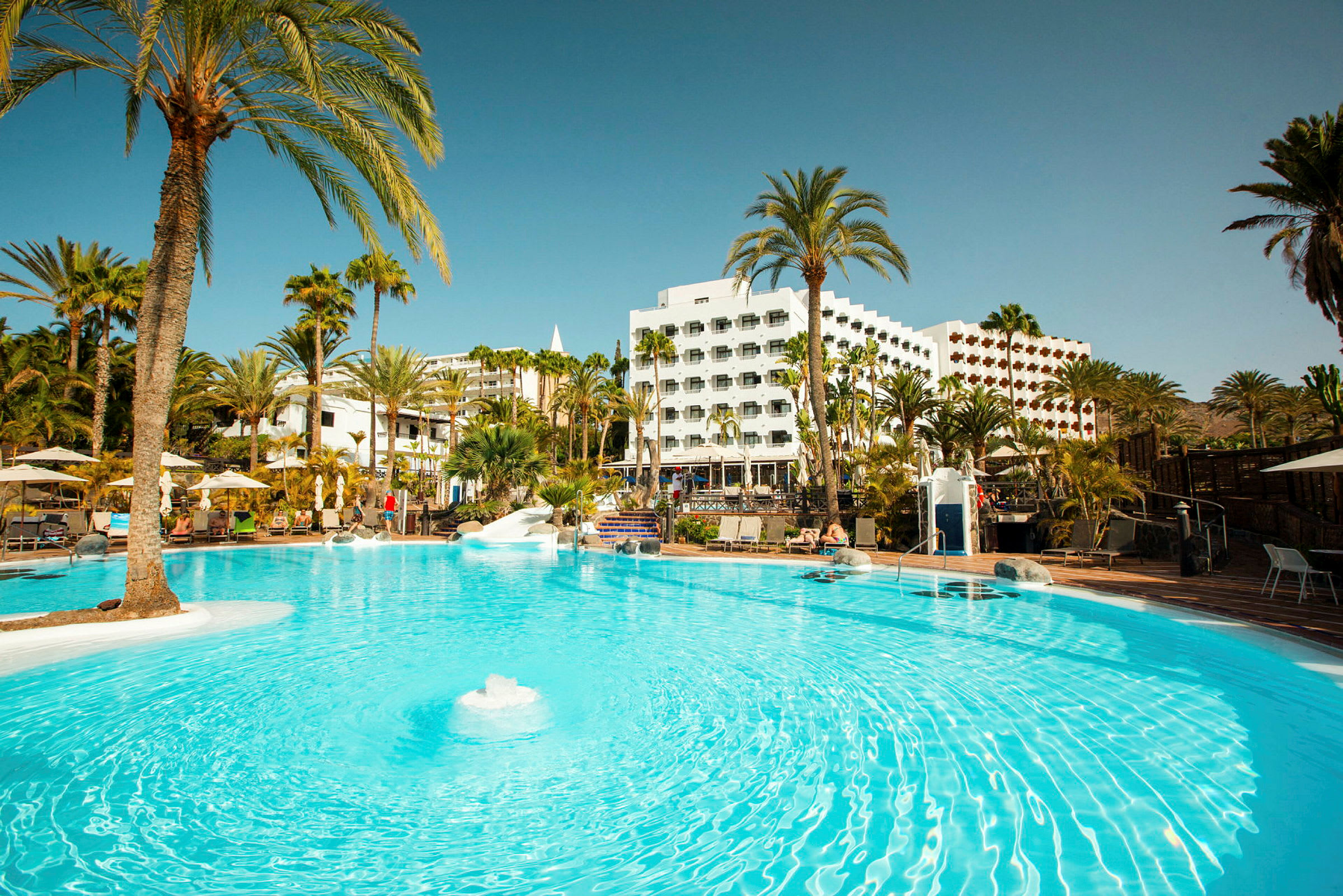 Billede af et hotelområde med udendørs pool og palmetræer. (AI-genereret tekst)