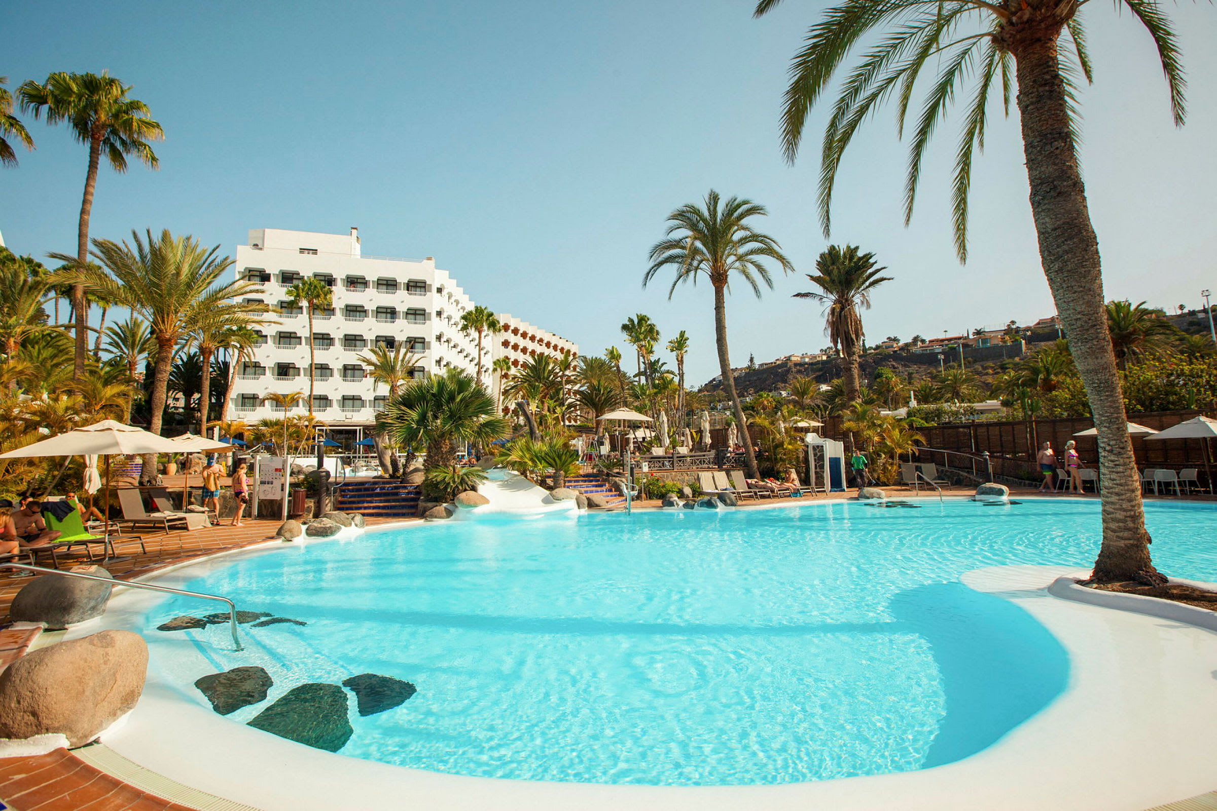 En swimmingpool omgivet af palmer og et hotel i baggrunden. (AI-genereret tekst)
