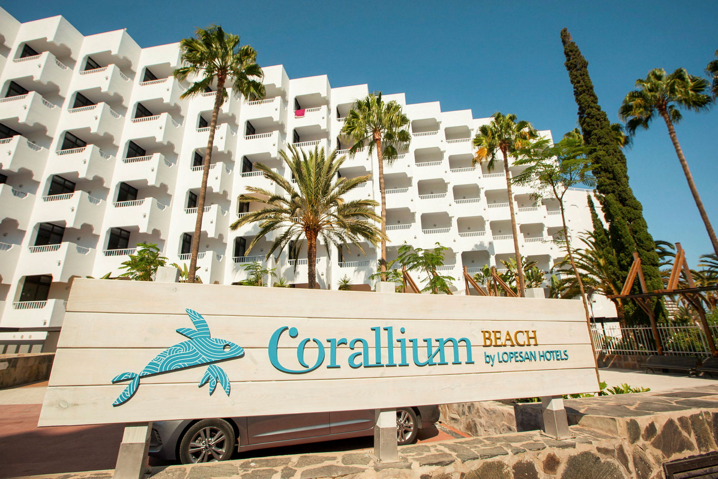 Hvid hotelbygning med altaner, omgivet af palmer, samt et skilt med teksten 'Corallium Beach by Lopesan Hotels'. (AI-genereret tekst)
