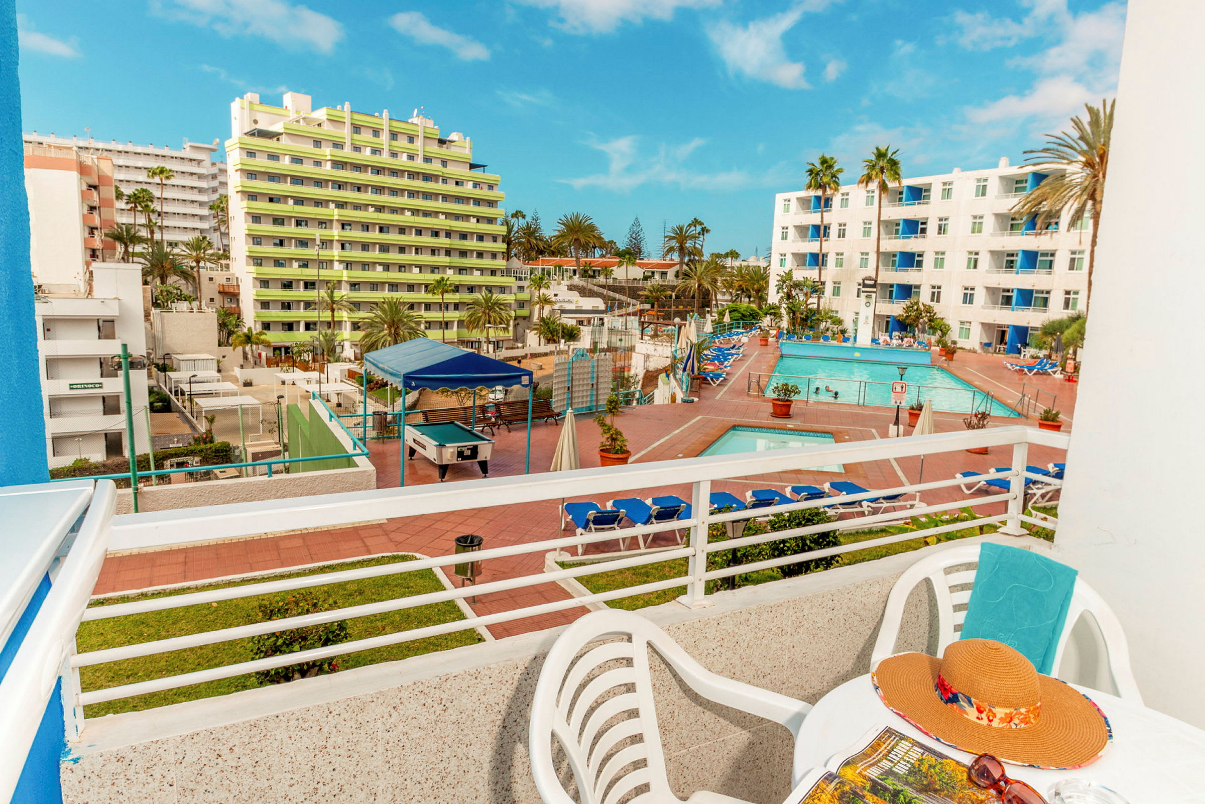 Udsigt fra en balkon, der viser et hotelområde med pool og liggestole samt bygninger i baggrunden. (AI-genereret tekst)