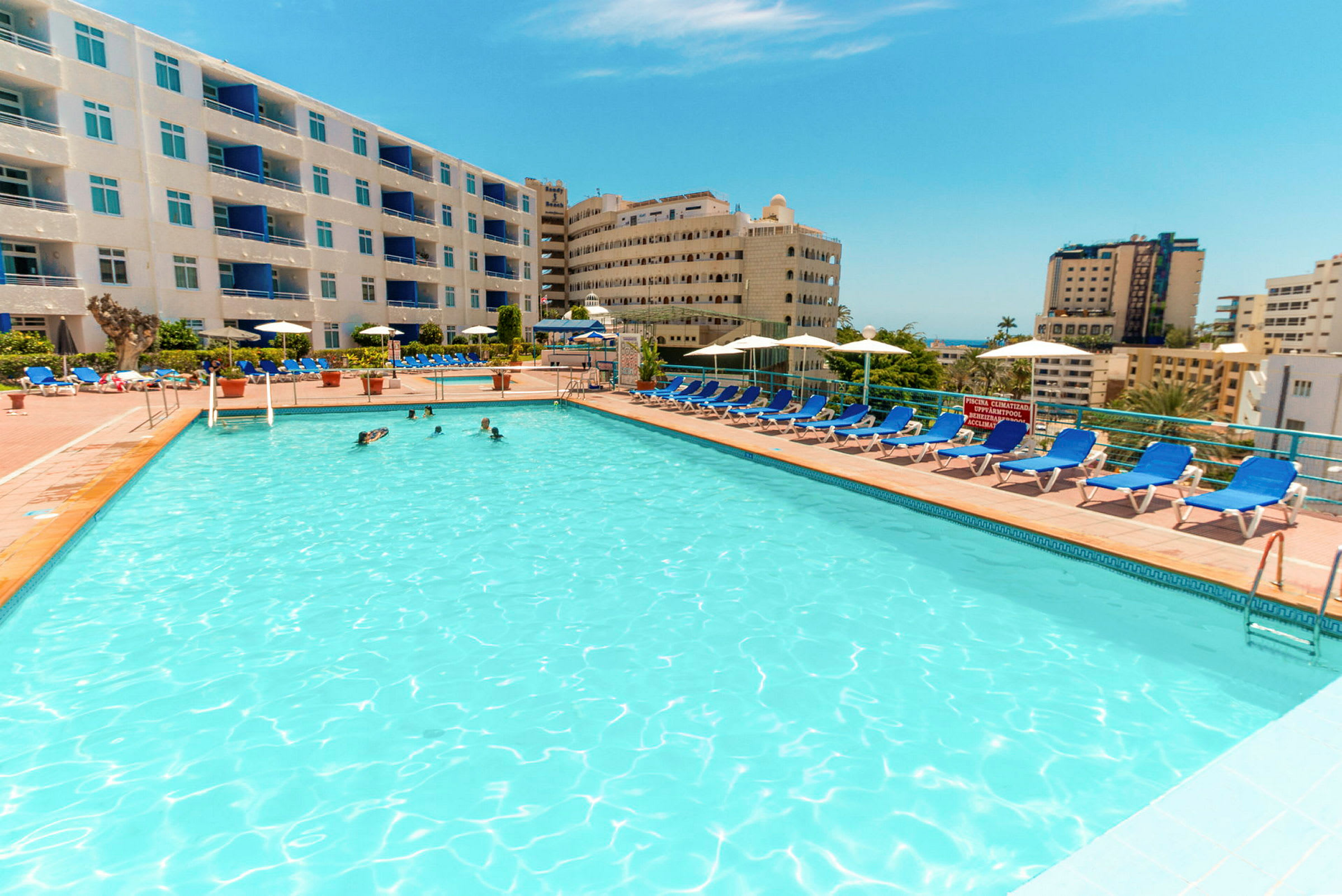 Stor udendørs swimmingpool omgivet af liggestole og hotelbygninger. (AI-genereret tekst)