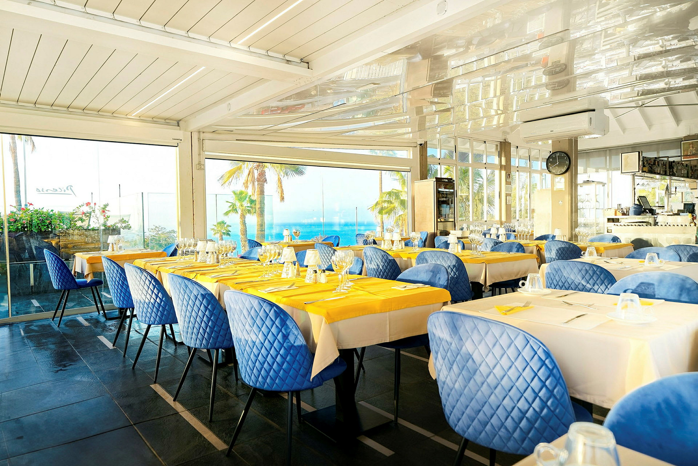 Restaurant med dækkede borde, blå stole og udsigt over palmer og havet gennem store vinduer. (AI-genereret tekst)