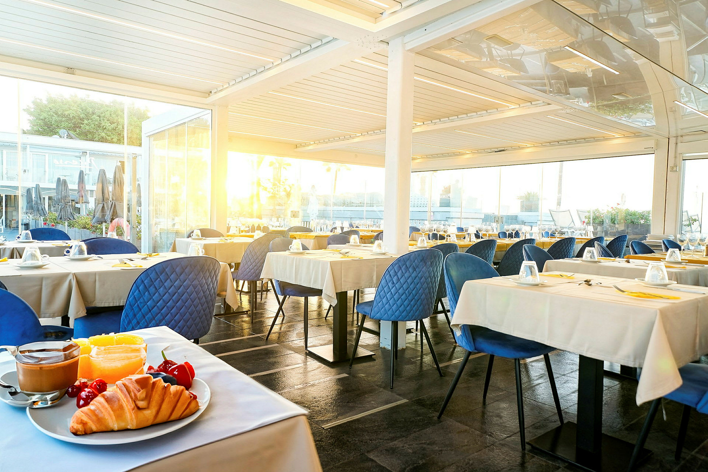 Billede af en restaurant med bordopsætning. Foran ses en tallerken med croissant, frugt og kaffe. (AI-genereret tekst)