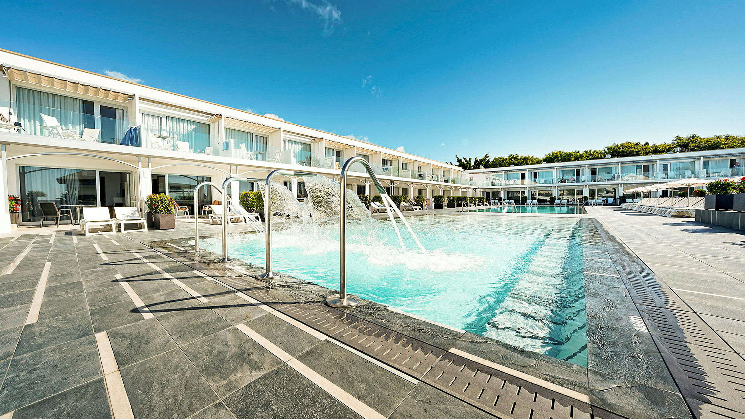 En udendørs pool med sprøjtefunktioner foran en række hotelværelser med terrasser. (AI-genereret tekst)