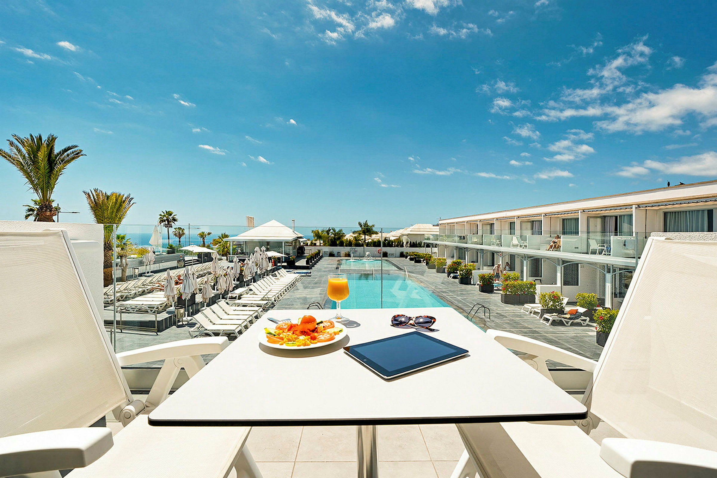 Udsigt fra en terrasse med bord og stole mod en pool og hotelbygning. (AI-genereret tekst)