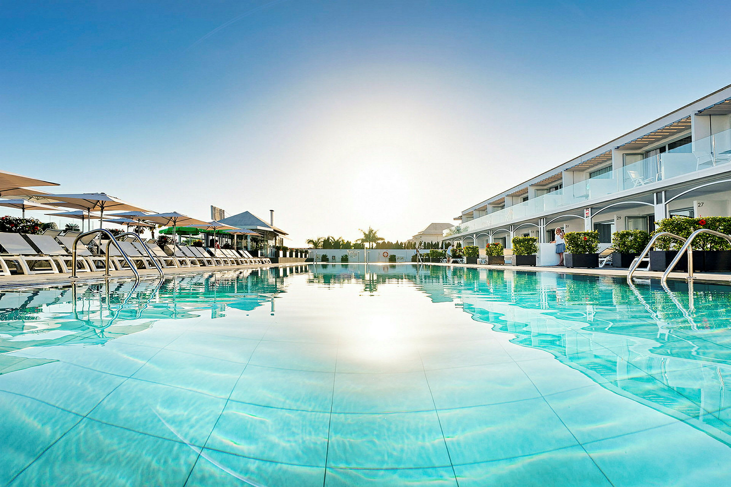Udsigt over en pool med liggestole og parasoller, omgivet af hotelbygninger. (AI-genereret tekst)