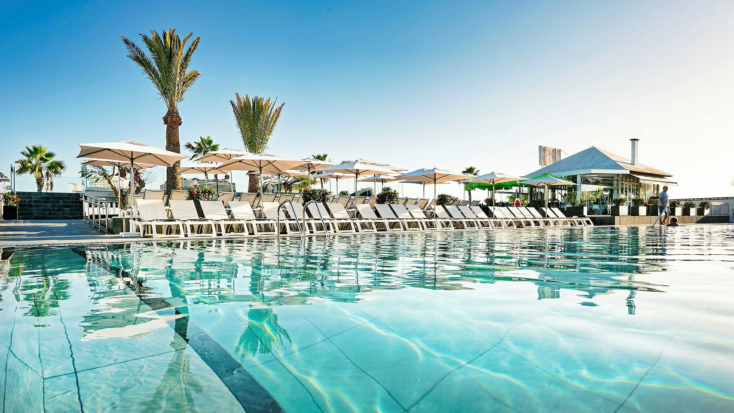 Billede af en pool med liggestole og parasoller ved et hotel. (AI-genereret tekst)