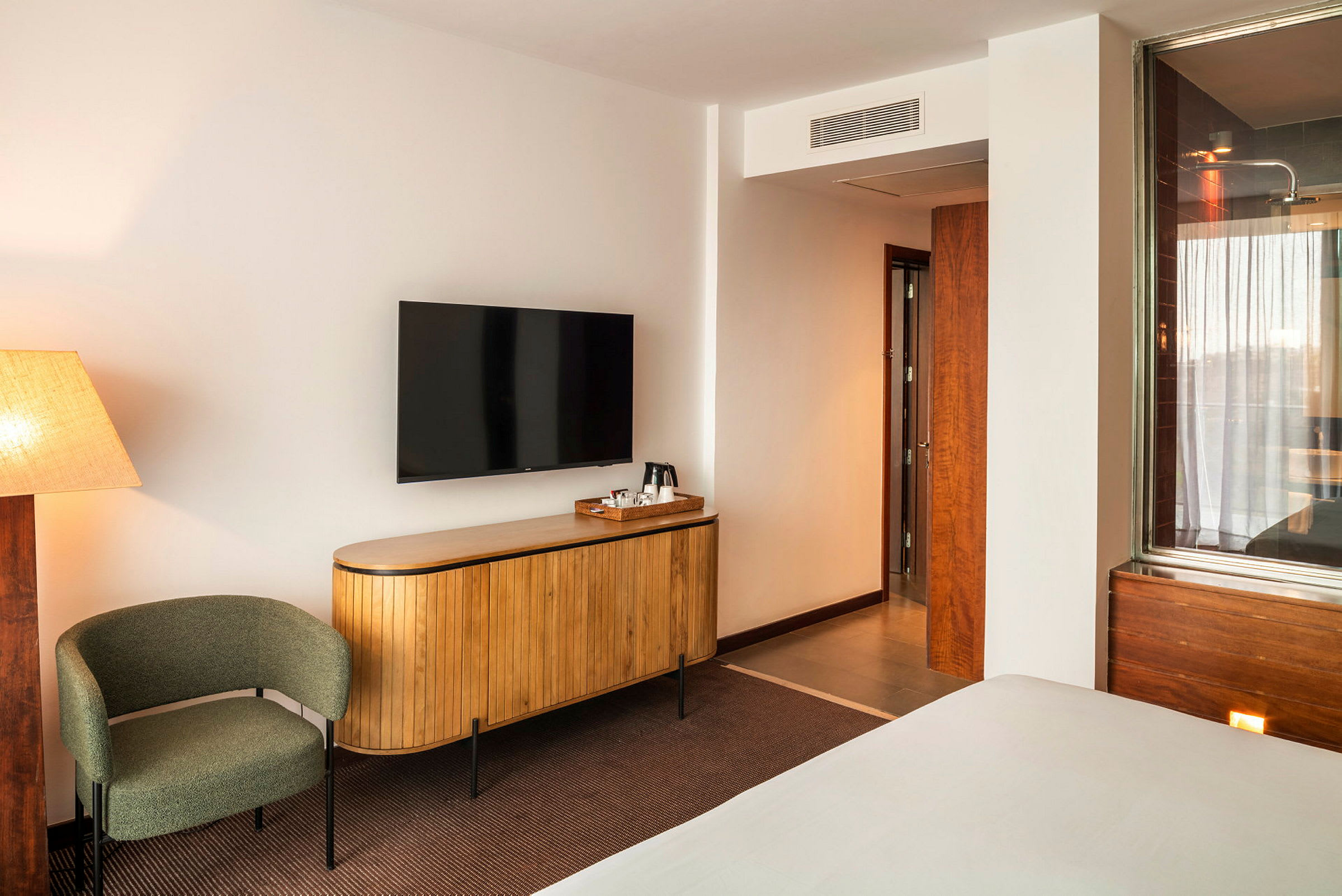 Et hotelværelse med beige vægge, en drejelig stol, et elegant tv monteret på væggen over en skænk i træ og en dør, der fører til badeværelset. (AI-genereret tekst)