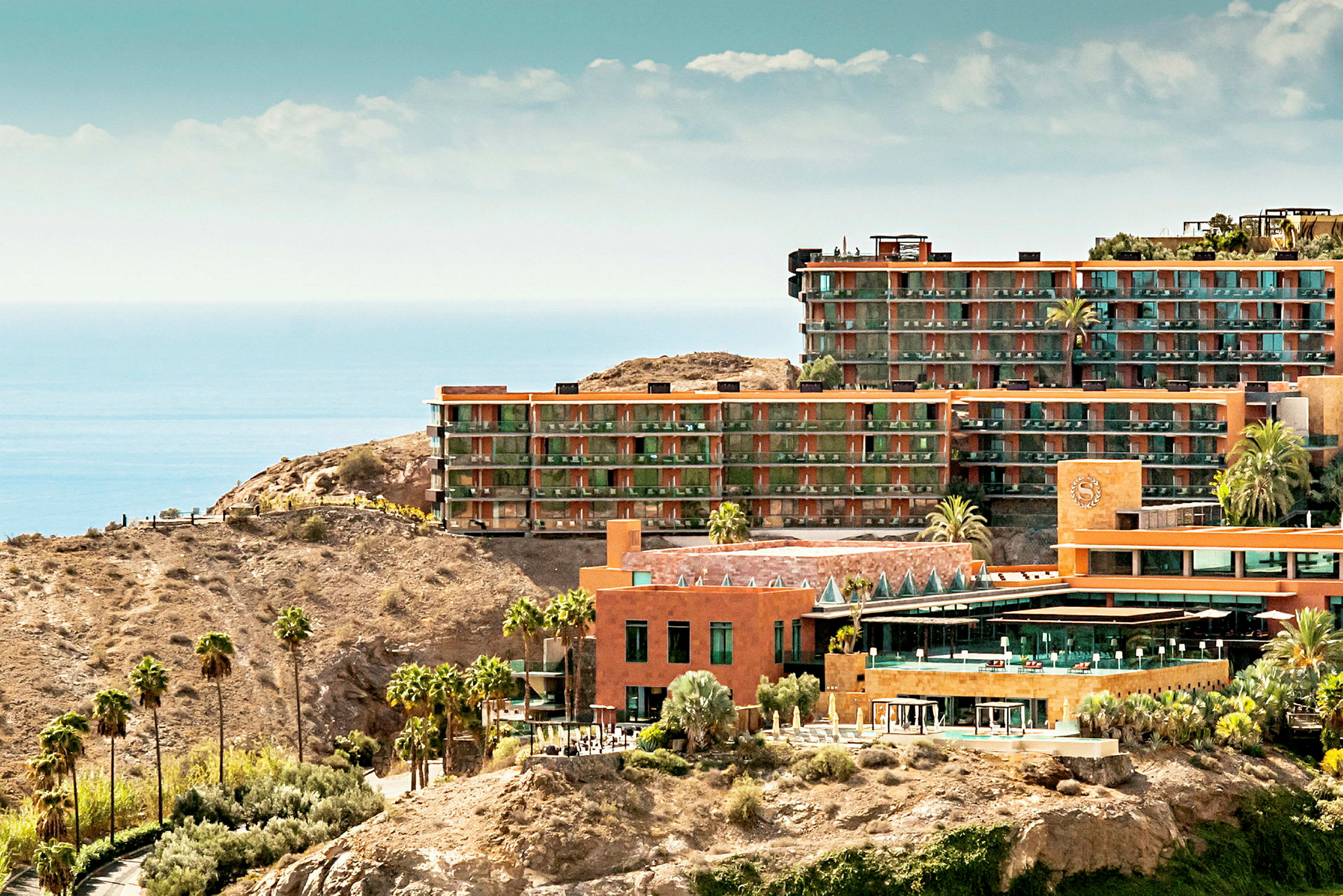 Billede af hotelbygninger i terrakotta-stil på klippeområde med udsigt over havet. (AI-genereret tekst)