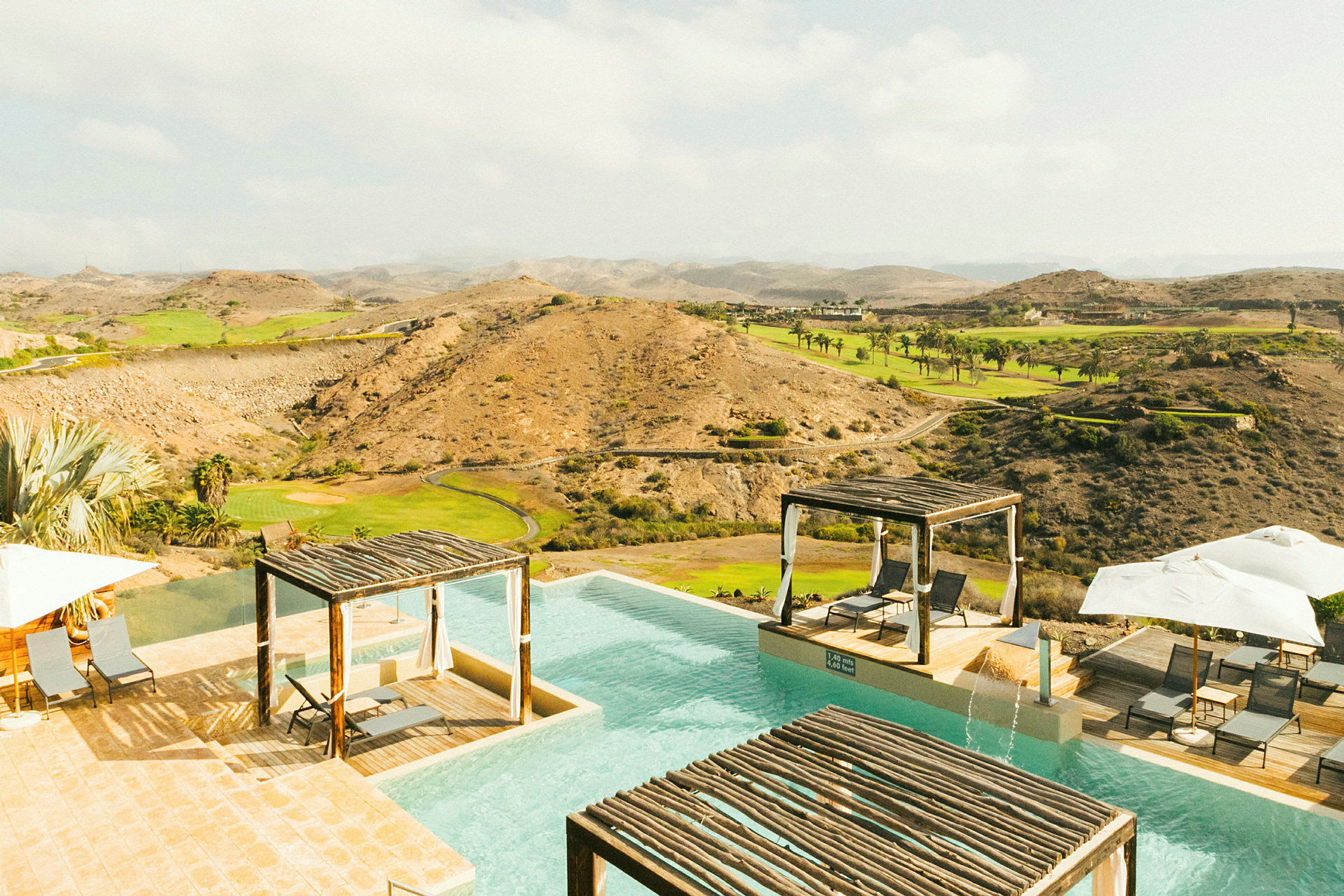 En infinity-pool med liggestole og pavilloner ved kanten, omgivet af et tørt landskab med grønne områder. (AI-genereret tekst)