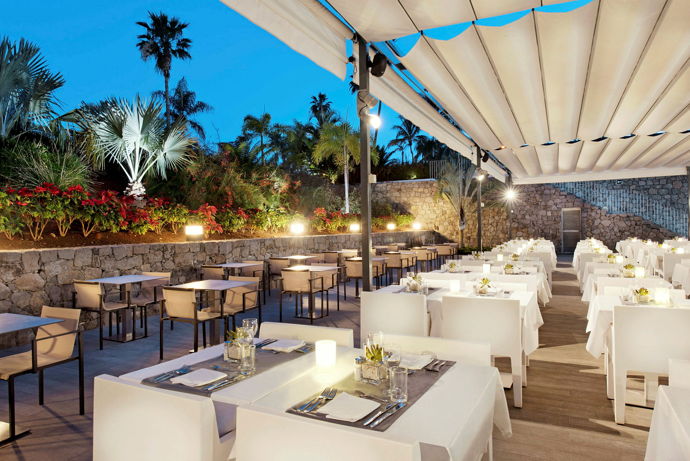 En udendørs restaurant med borde og stole under en stor markise. Blomstrende planter og palmer ses ved siden af stenmurerne. (AI-genereret tekst)