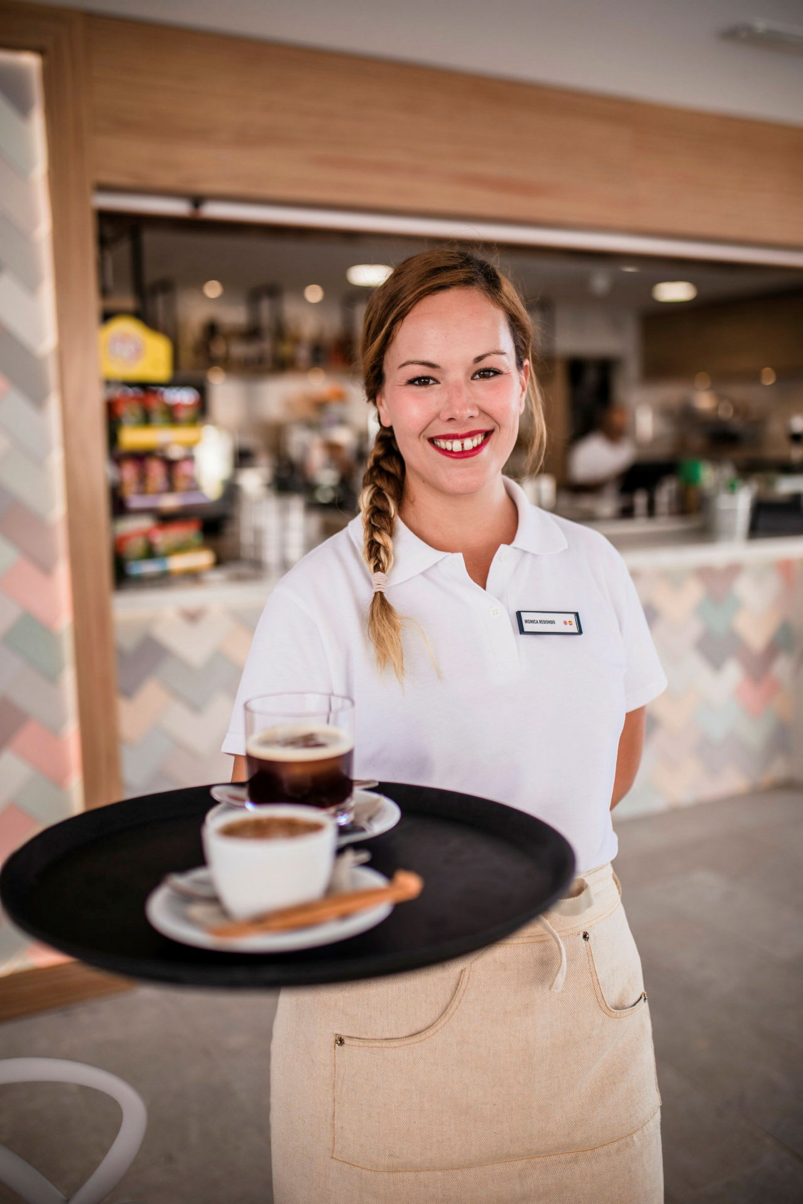 En servitrice i uniform holder en bakke med kaffe og te foran en baggrund med en cafédisk. (AI-genereret tekst)