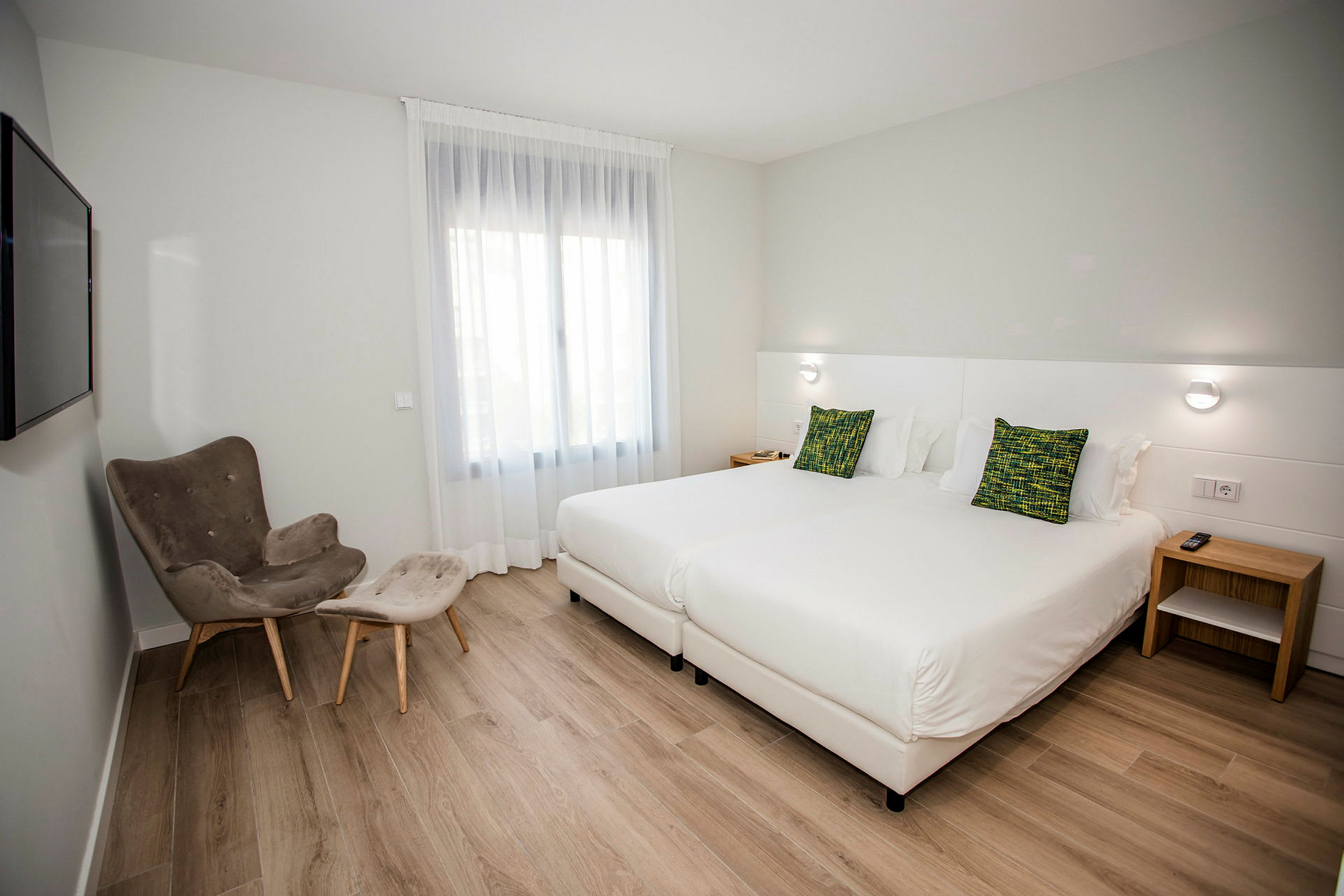 Hotelværelse med to enkeltsenge, en lænestol med fodskammel og et vindue med lyst stof. (AI-genereret tekst)