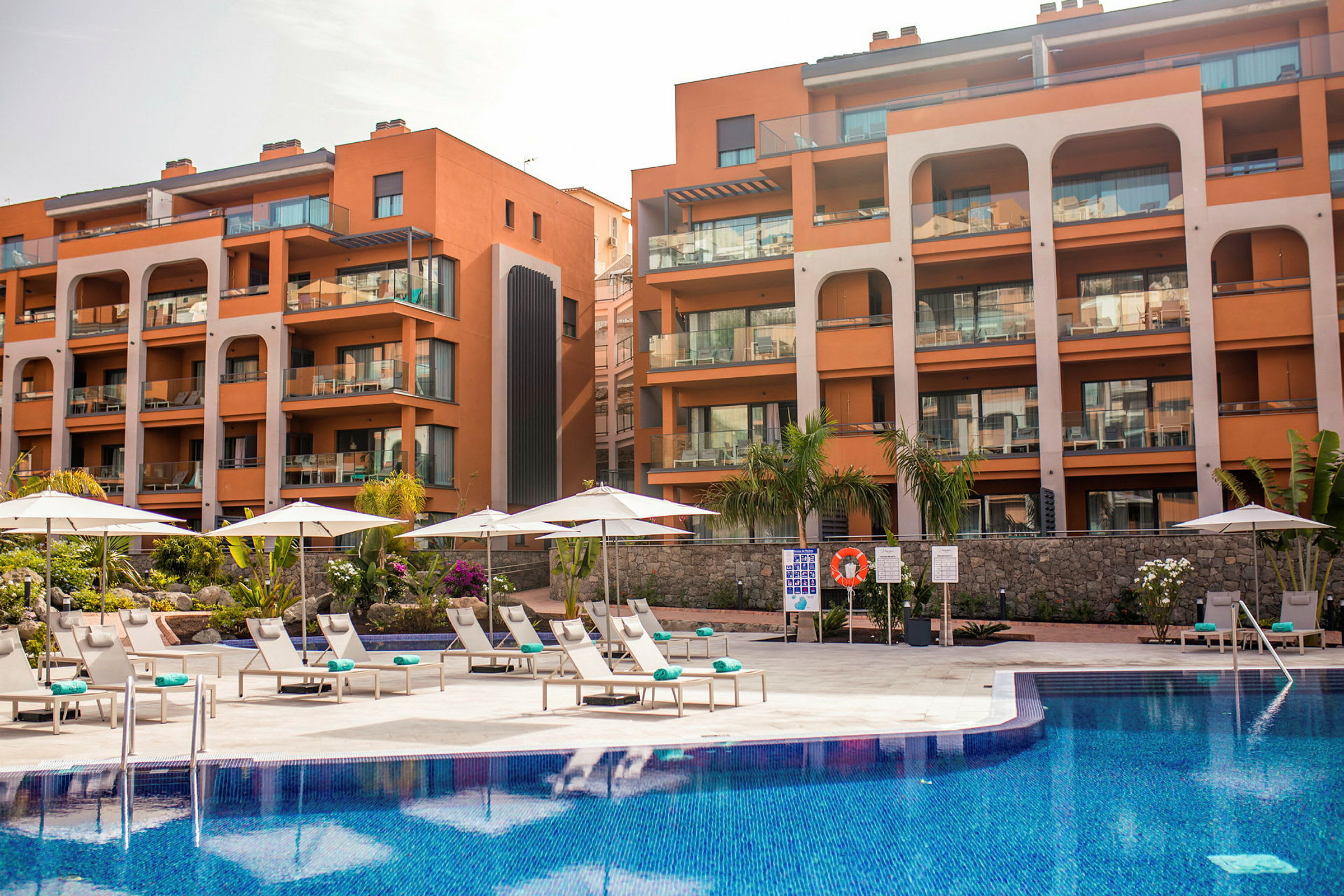 Hotelbygning med flere altaner og en pool foran med solsenge og parasoller spredt ud. (AI-genereret tekst)
