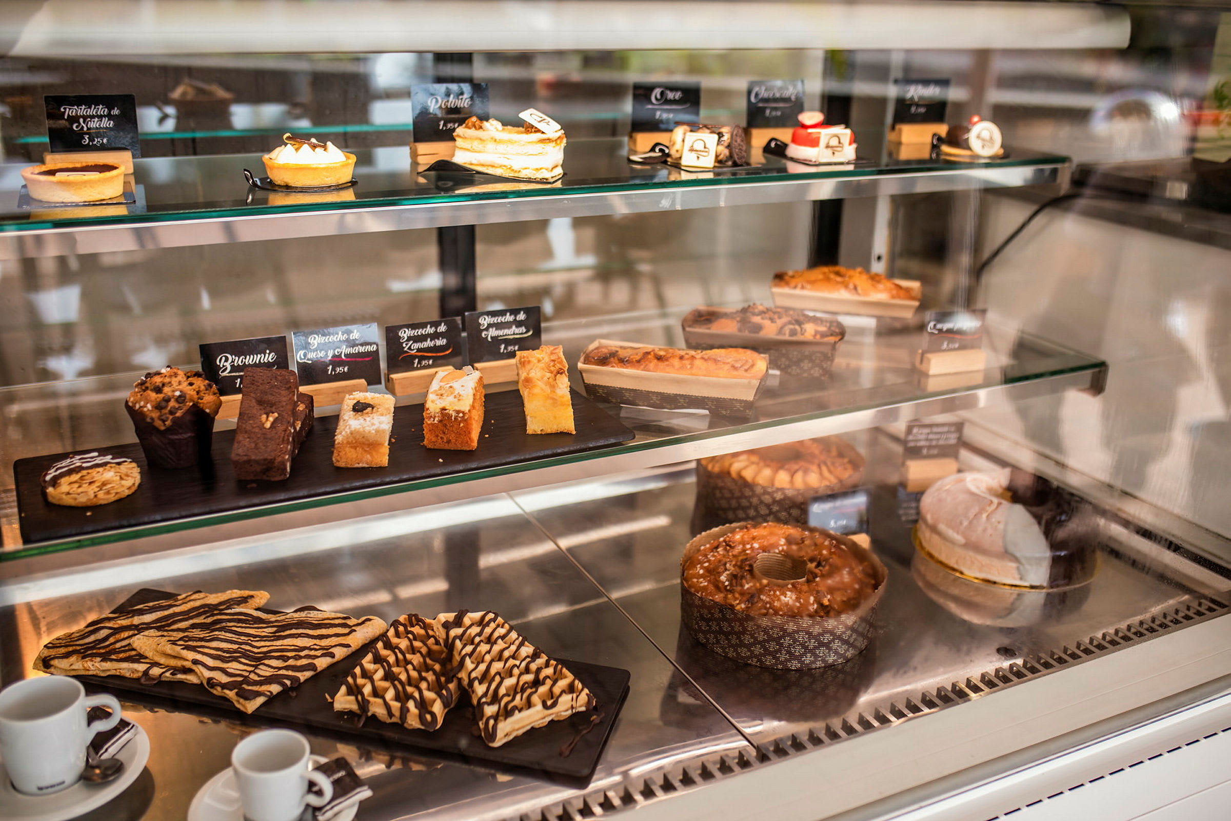 Et køleskab med forskellige bagværk og desserter, herunder muffins, kager og tærter, opstillet på hylder. (AI-genereret tekst)