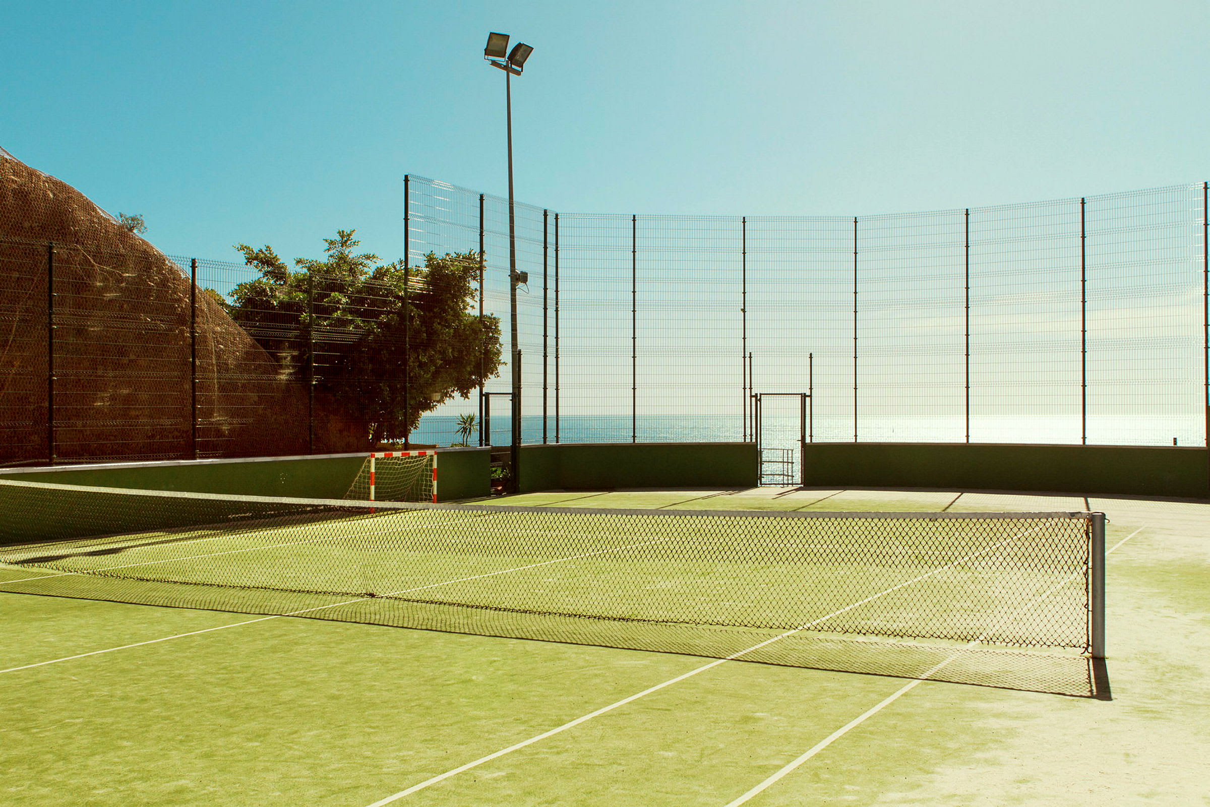 En tennisbane omgivet af nethegn nær havet. (AI-genereret tekst)
