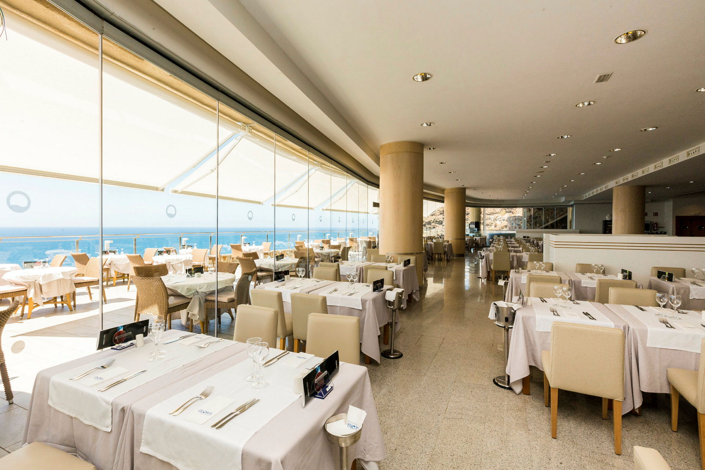 En restaurant med borde dækket med hvide duge og glas, med udsigt over havet gennem store vinduer. (AI-genereret tekst)
