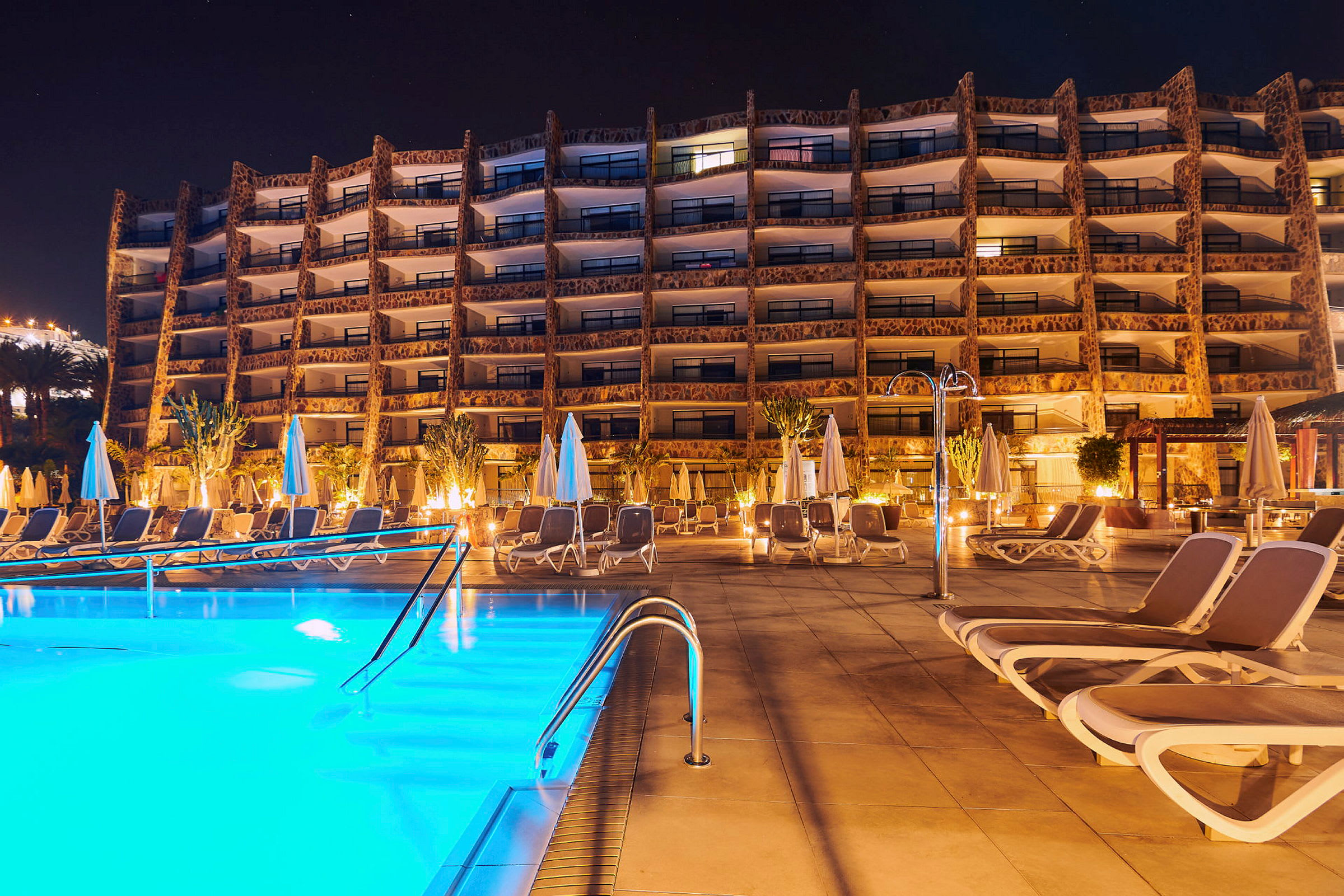 Billede af hotelbygning med pool og liggestole i forgrunden, fotograferet om aftenen. (AI-genereret tekst)