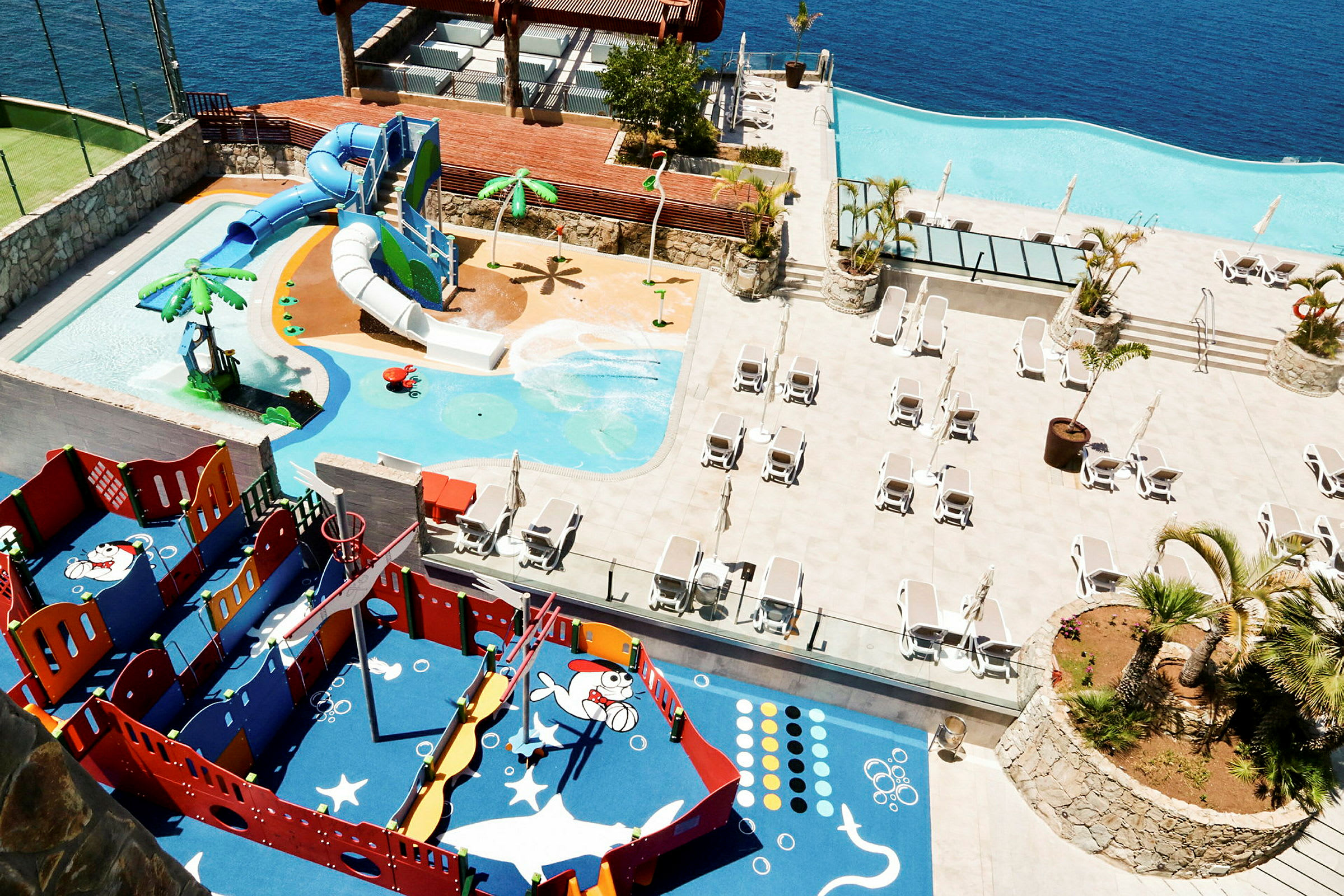 Børnepool med vandrutsjebaner og legeplads ved siden af liggestole og infinity-pool. (AI-genereret tekst)