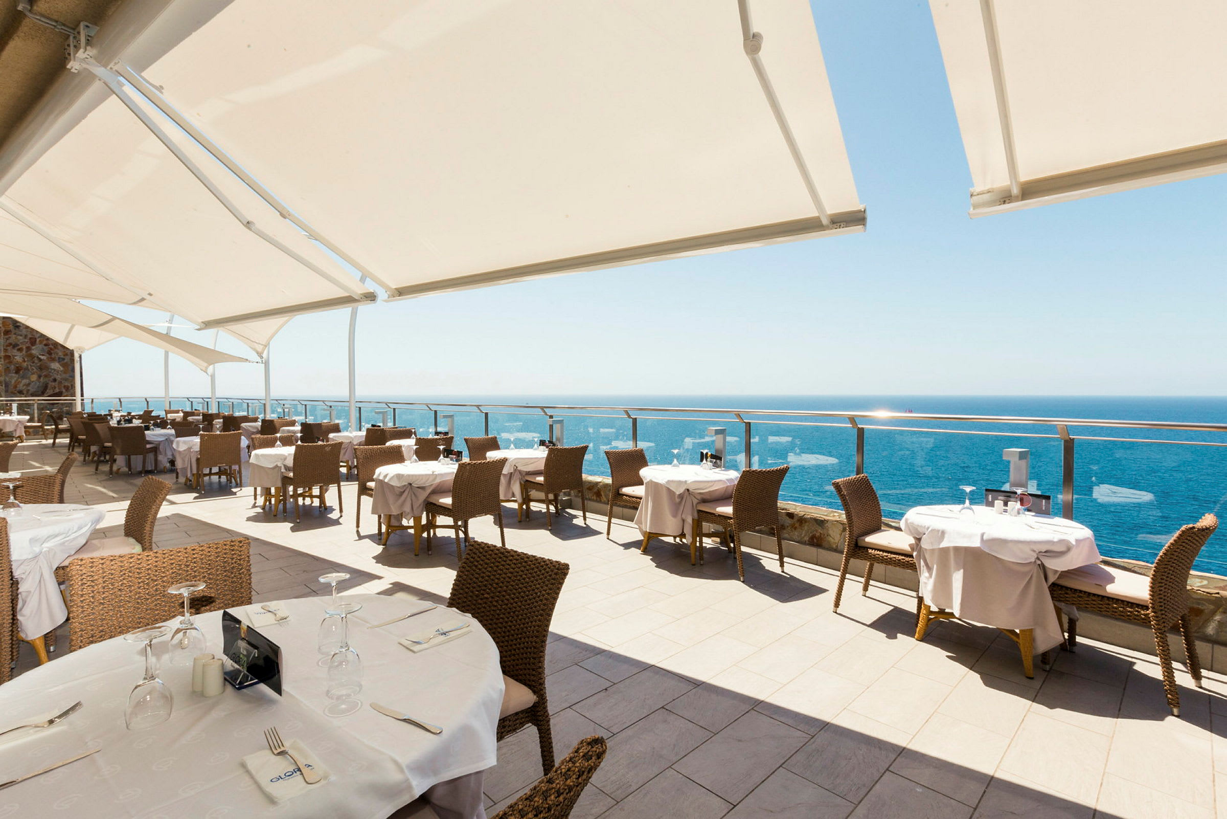 En udendørs restaurant med havudsigt og solbeskyttelse over siddepladserne. (AI-genereret tekst)
