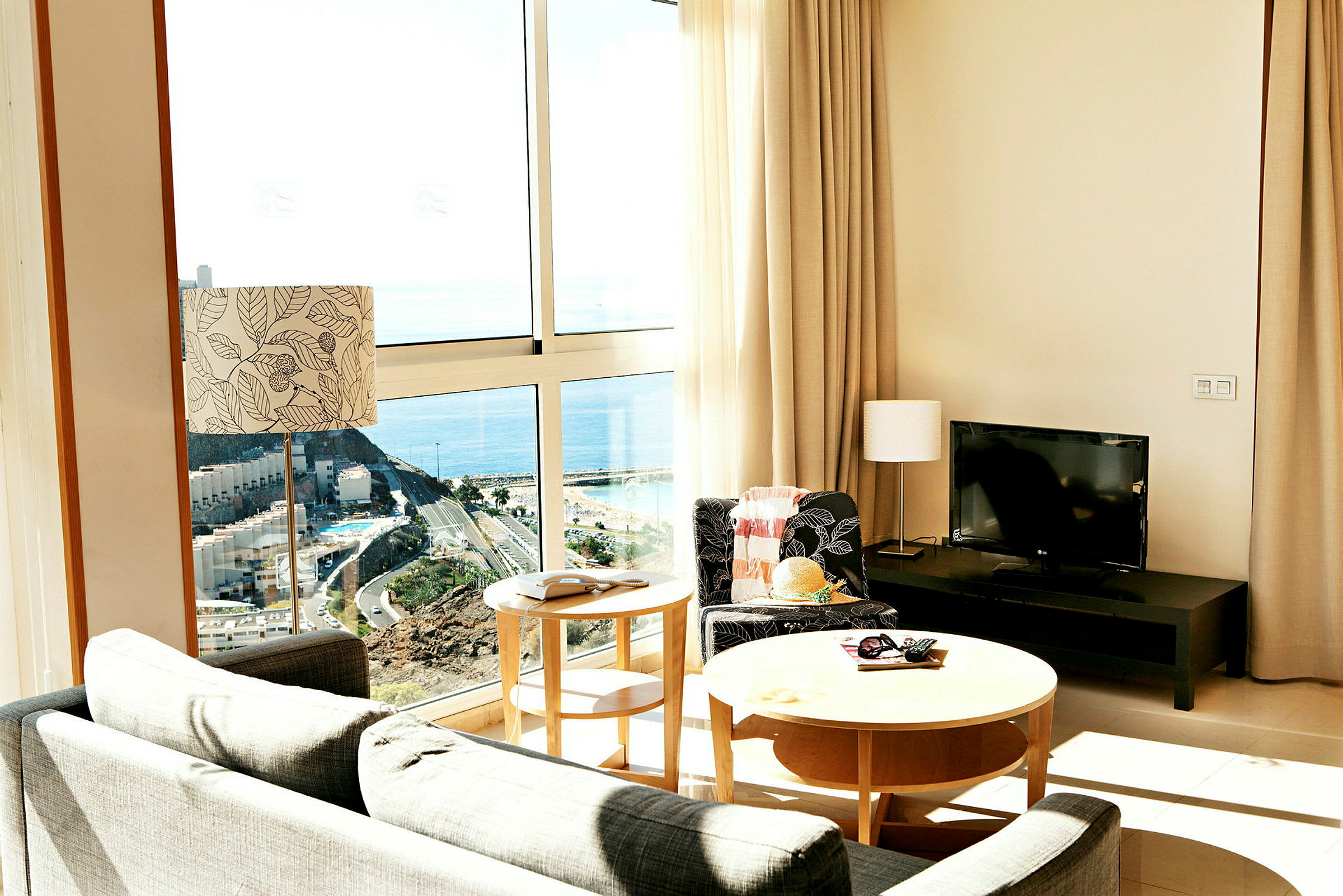 Interiør af et rum med store vinduer og udsigt til havet. Møbler inkluderer sofa, bord og en tv. (AI-genereret tekst)