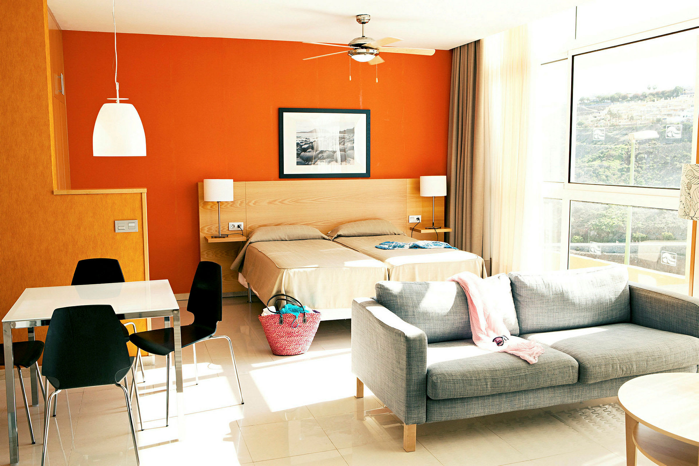 Billede af et hotelværelse med to enkeltsenge, grå sofa og et spisebord med sorte stole. Væggene er orange og store vinduer lader lys komme ind. (AI-genereret tekst)
